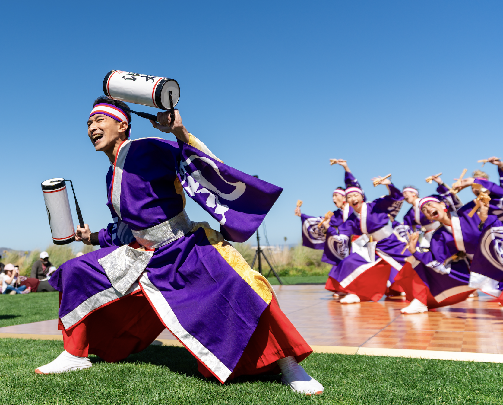 Japanese festival dancers in colorful purple costumes performing with drums outdoors under a clear blue sky