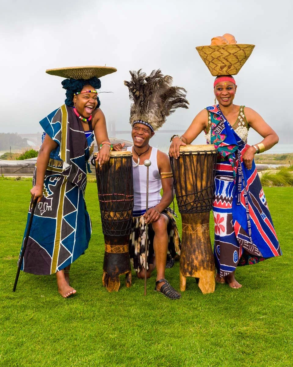 Three African performers in colorful traditional clothing and headdresses pose with tall hand drums on a grassy waterfront lawn.