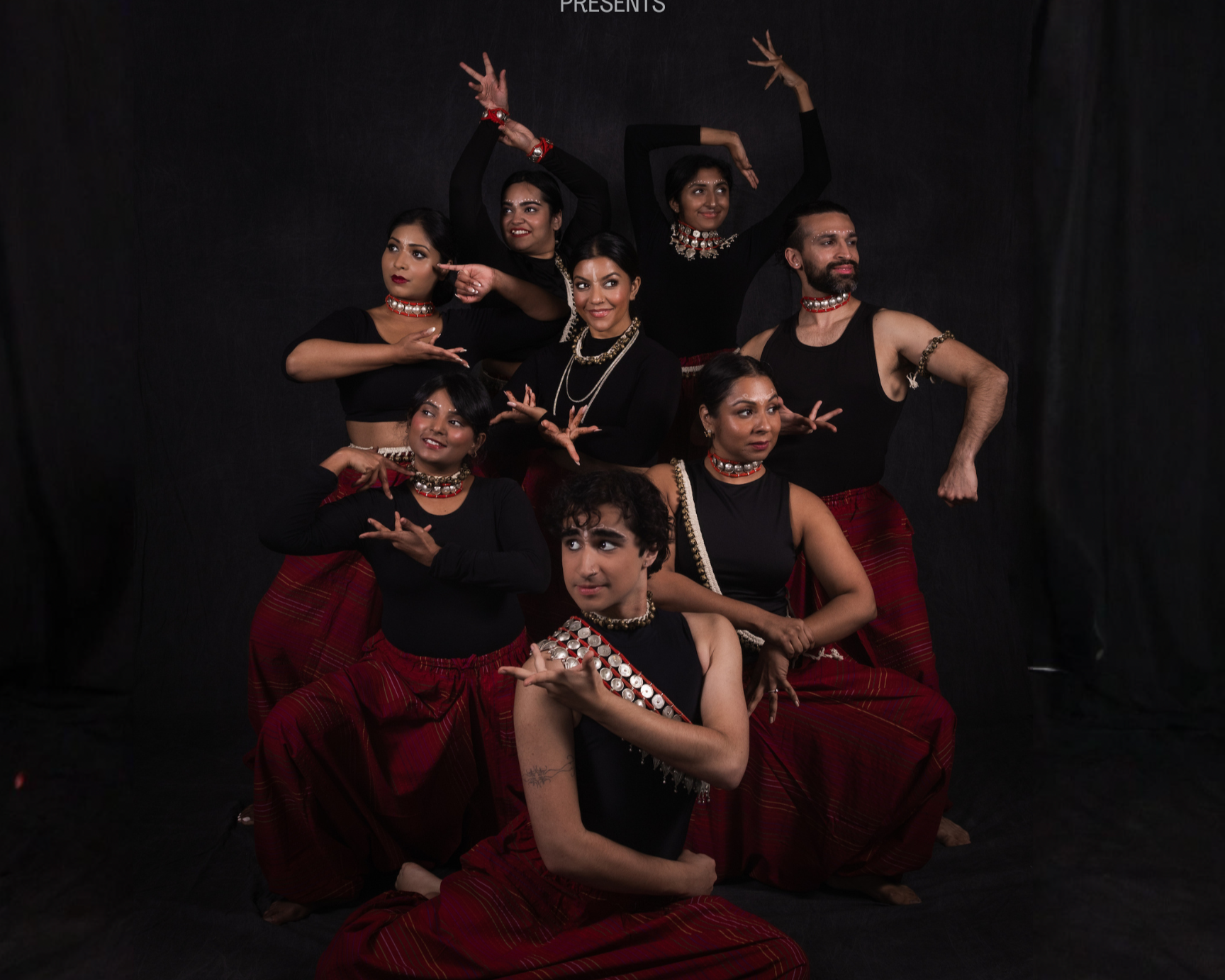 Group of South Asian dancers in black tops and red skirts posing in expressive choreography against a black backdrop.