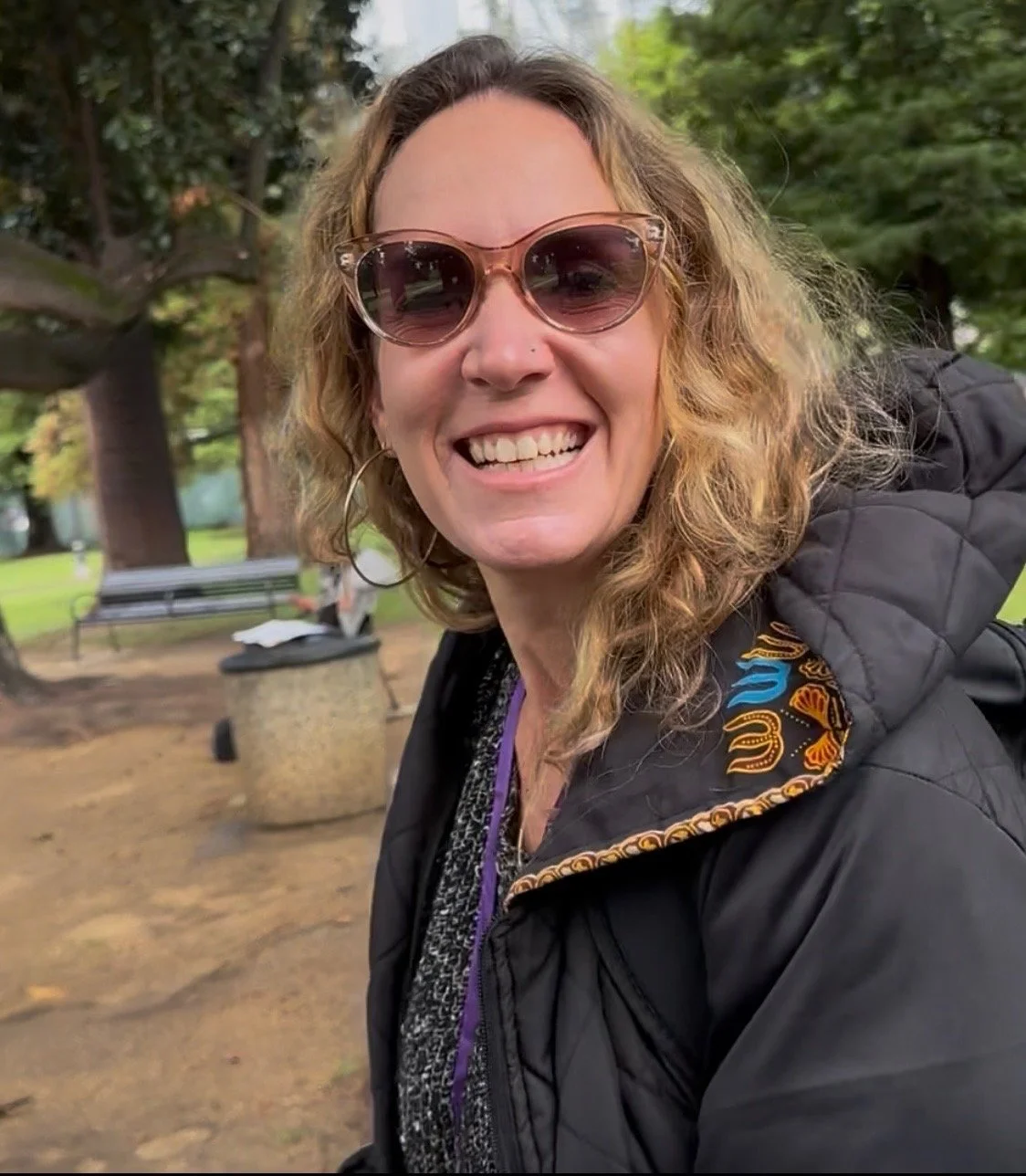 Smiling woman wearing sunglasses and a black jacket outdoors in a park