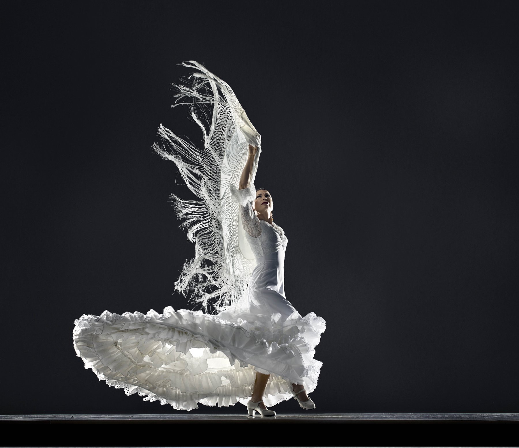 Flamenco dancer in a flowing white ruffled dress and lace shawl performing against a black background