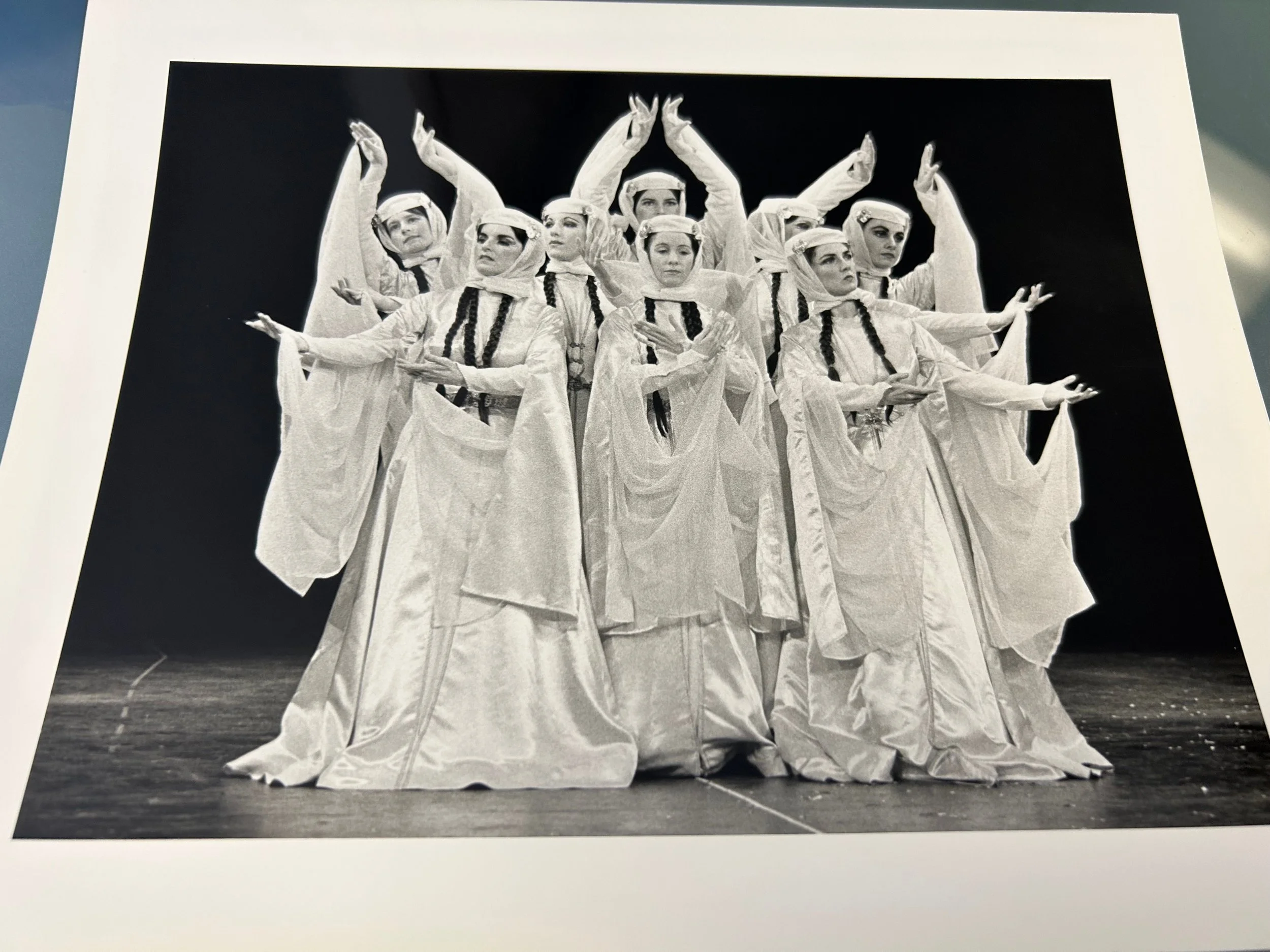 Black-and-white photo of a ballet ensemble in flowing white costumes performing synchronized poses on stage.