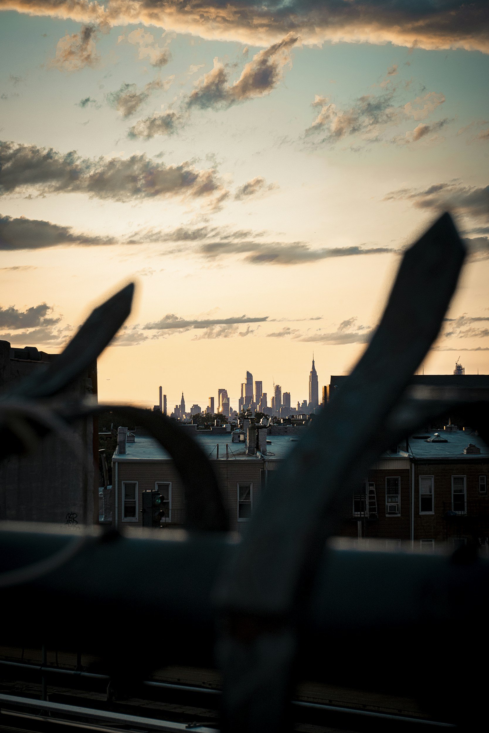 Sunset over the New York City skyline viewed through a rooftop fence, with clouds in the sky and silhouette of buildings including the Empire State Building.