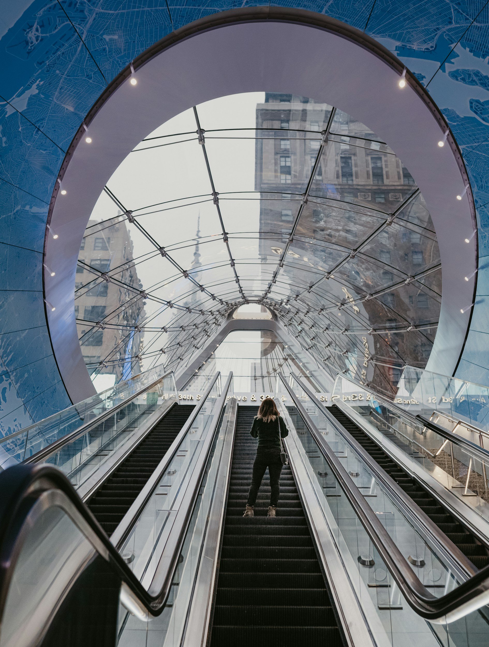 A person standing on an escalator inside a modern building with a large circular glass ceiling reflecting city buildings.