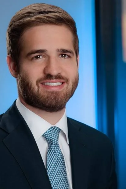 A man with brown hair and a beard, wearing a dark suit, white shirt, and a blue patterned tie, smiling in front of a blue background.