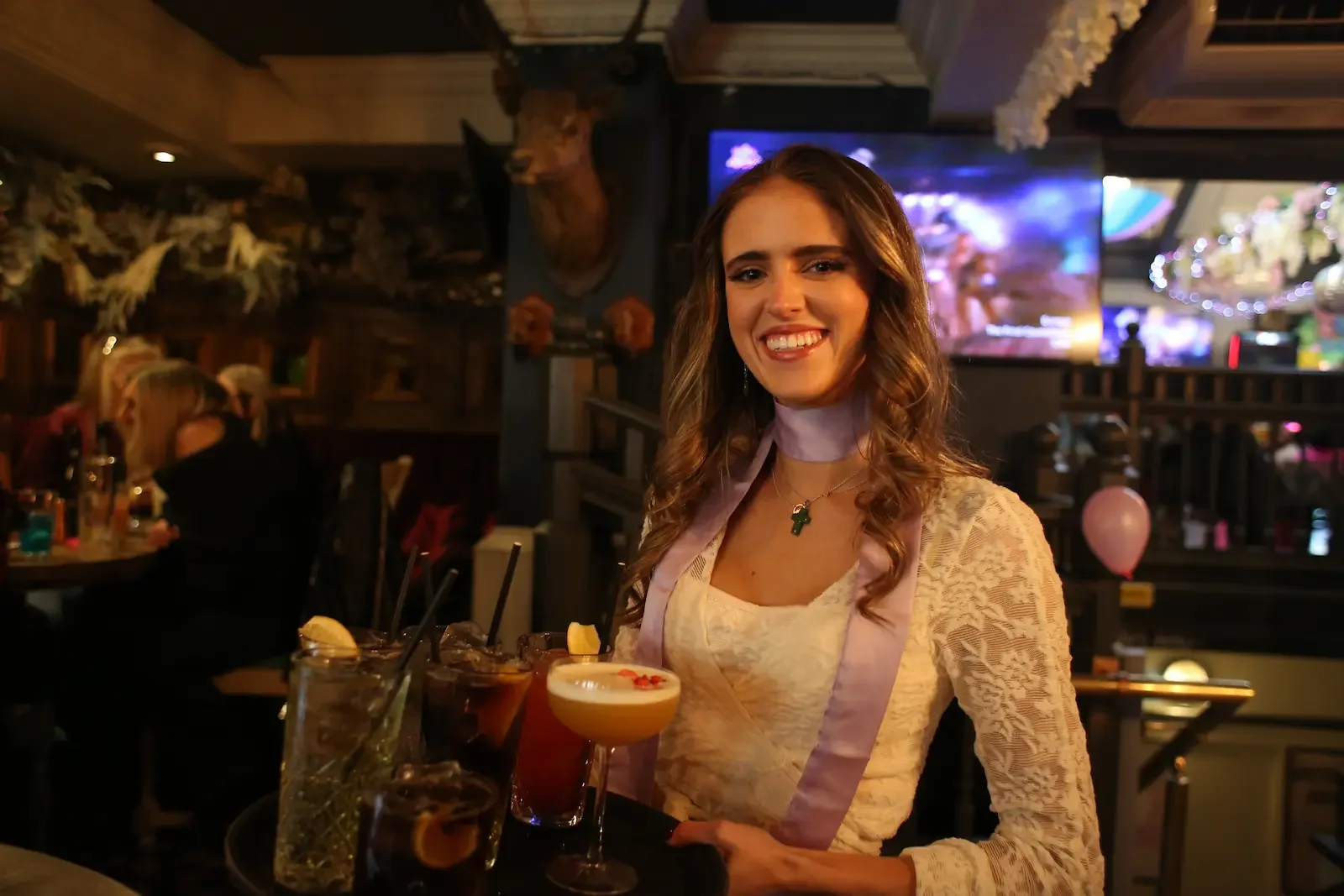 A young woman smiling and sitting at a table in a bar or restaurant, surrounded by drinks, with a television screen and other patrons in the background.