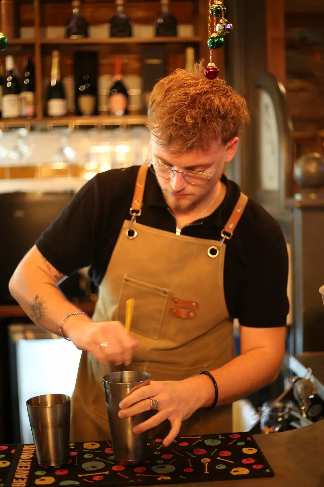 A bartender with glasses and tattoos on his arm, wearing a black shirt and a tan apron, prepares a drink at a bar with bottles and glasses in the background.