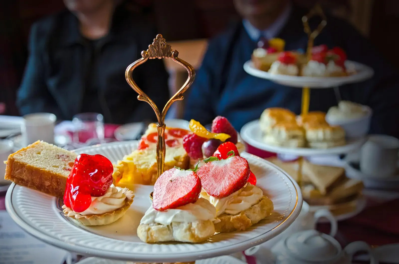 A close-up of a dessert platter featuring strawberries, raspberries, and a piece of cake with a dollop of whipped cream, on a fancy white plate with a gold trim, in a setting with tiered dessert stands and people in the background.