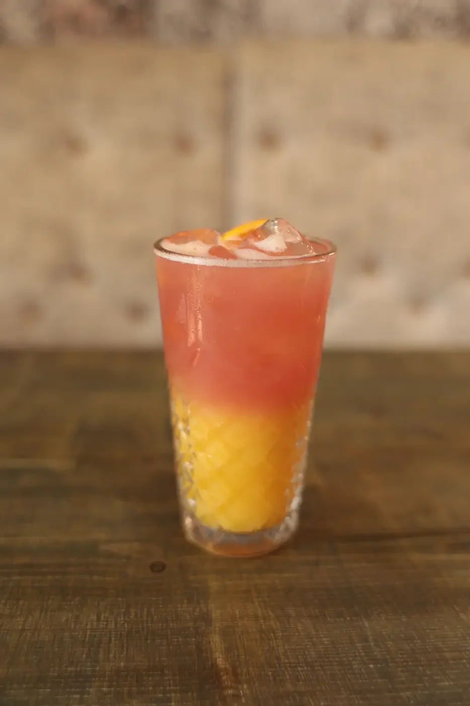 A colorful cocktail in a tall glass with a yellow fruit garnish, placed on a wooden table with a blurred background.