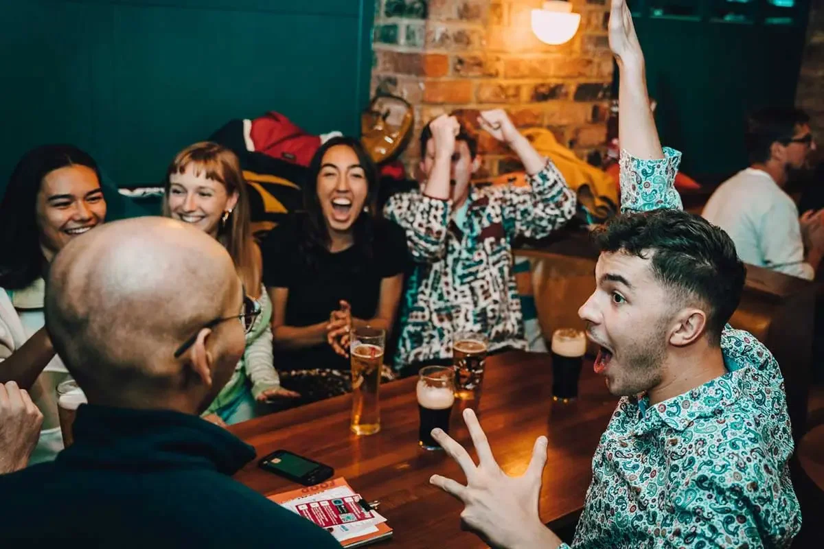 Group of friends at a table in a bar, celebrating and having fun, with drinks and lively expressions.