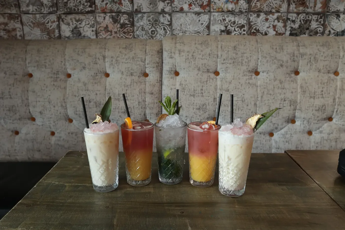 Five colorful cocktails on a wooden table with a beige upholstered booth in the background. The drinks have garnishes such as pineapple, mint, and fruit slices.