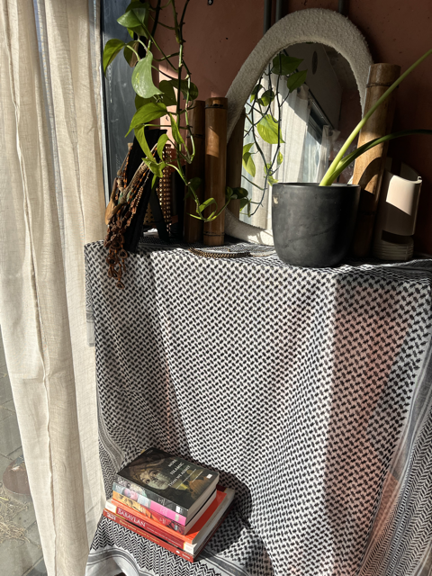A dresser covered with a keffiyeh, with stacked books on the floor, a mirror, potted plants, and some decorative objects on top.