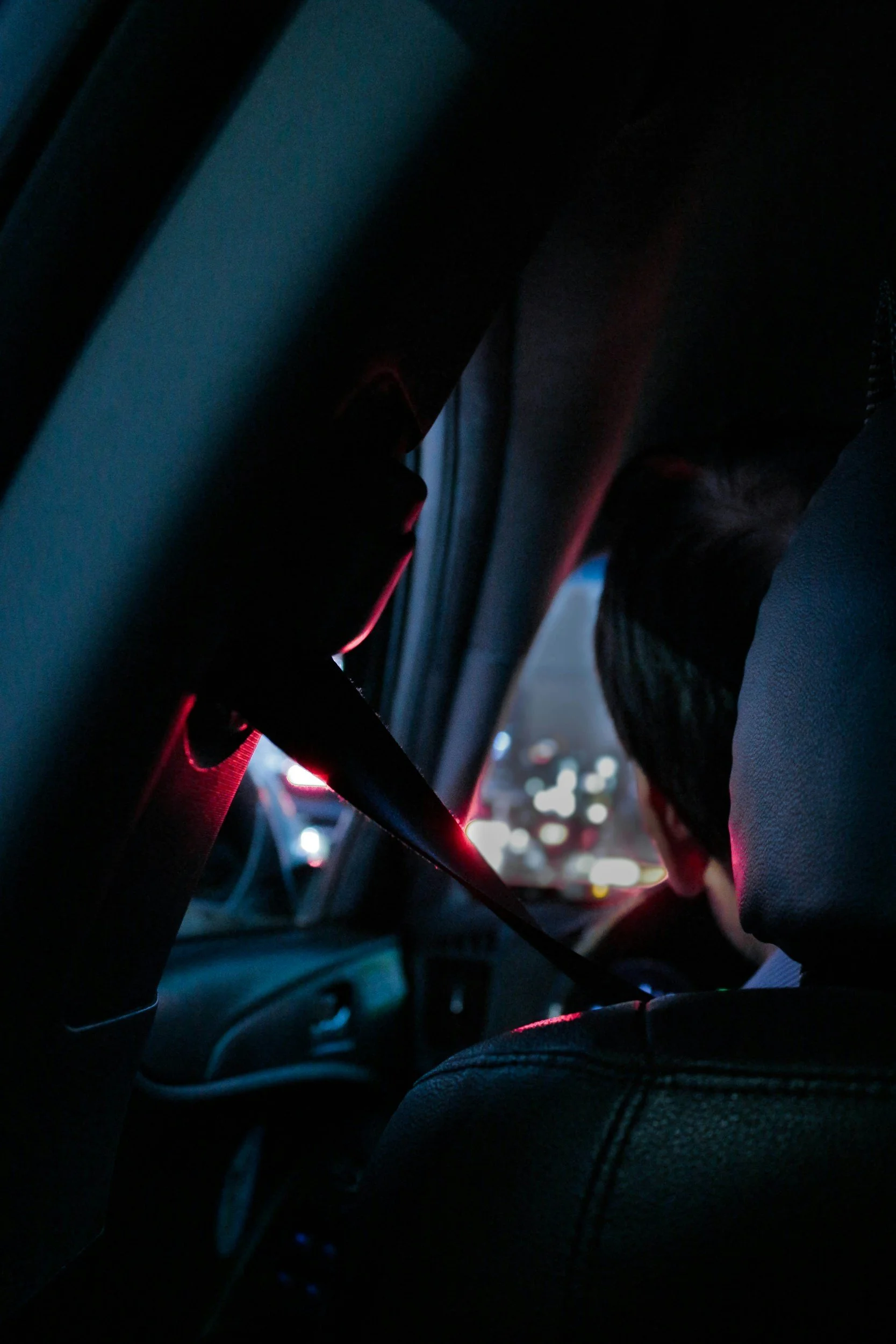 Photo of the inside of a car from the back seat. There is someone driving at night there are read lights present.