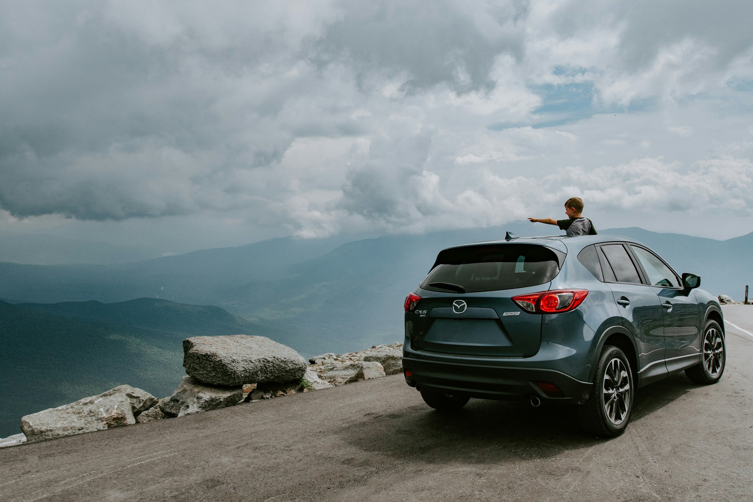 A photo of a grey mazda with a family on a mountaintop. There is a child pointing out the sunroof.