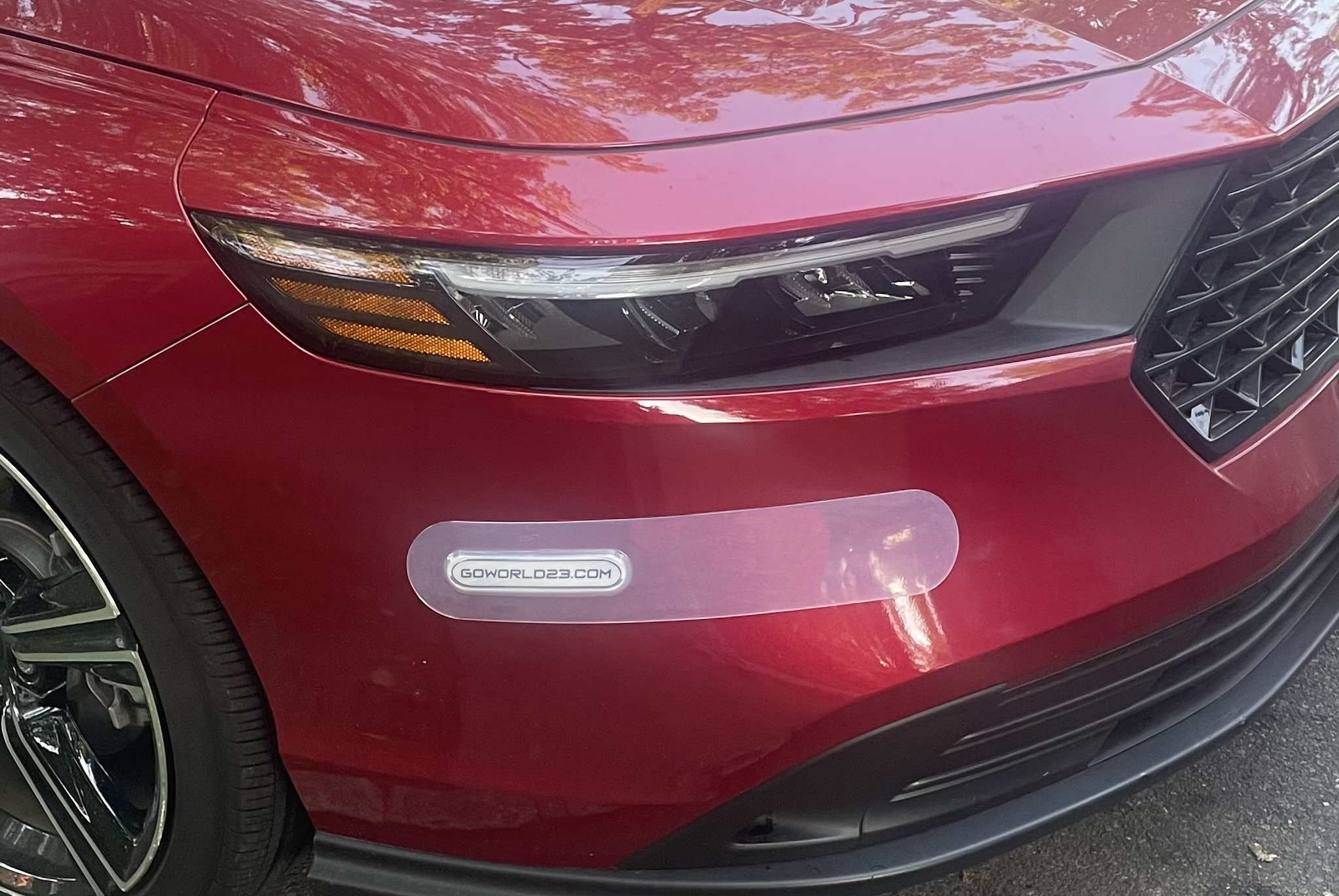 Close-up of the front end of a red car with a headlight, a front grille, and a reflective sticker with the text 'GOWORLD23.COM' on the front bumper.