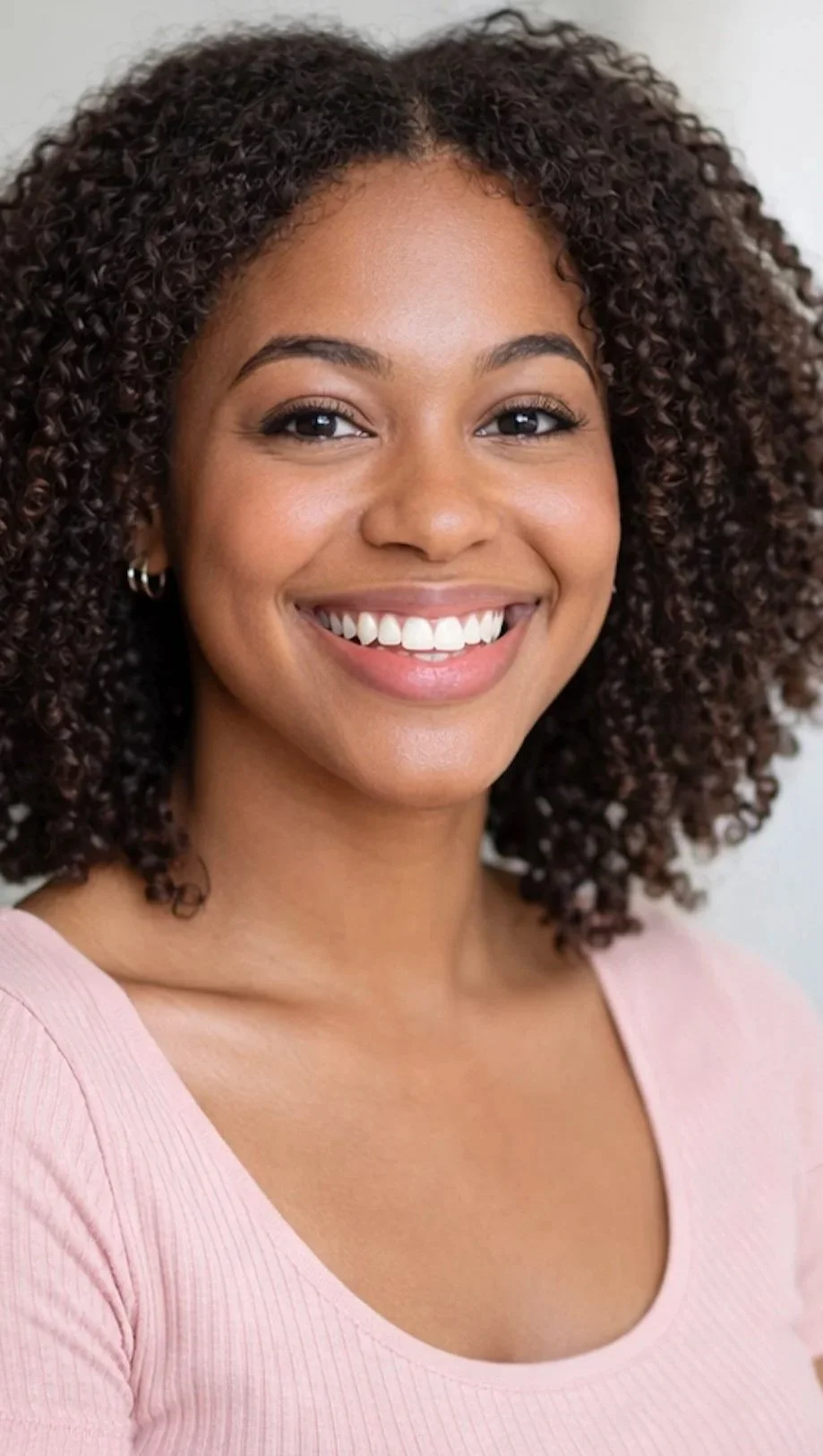 A woman with curly black hair, and pink blouse, smiling in an indoor setting.