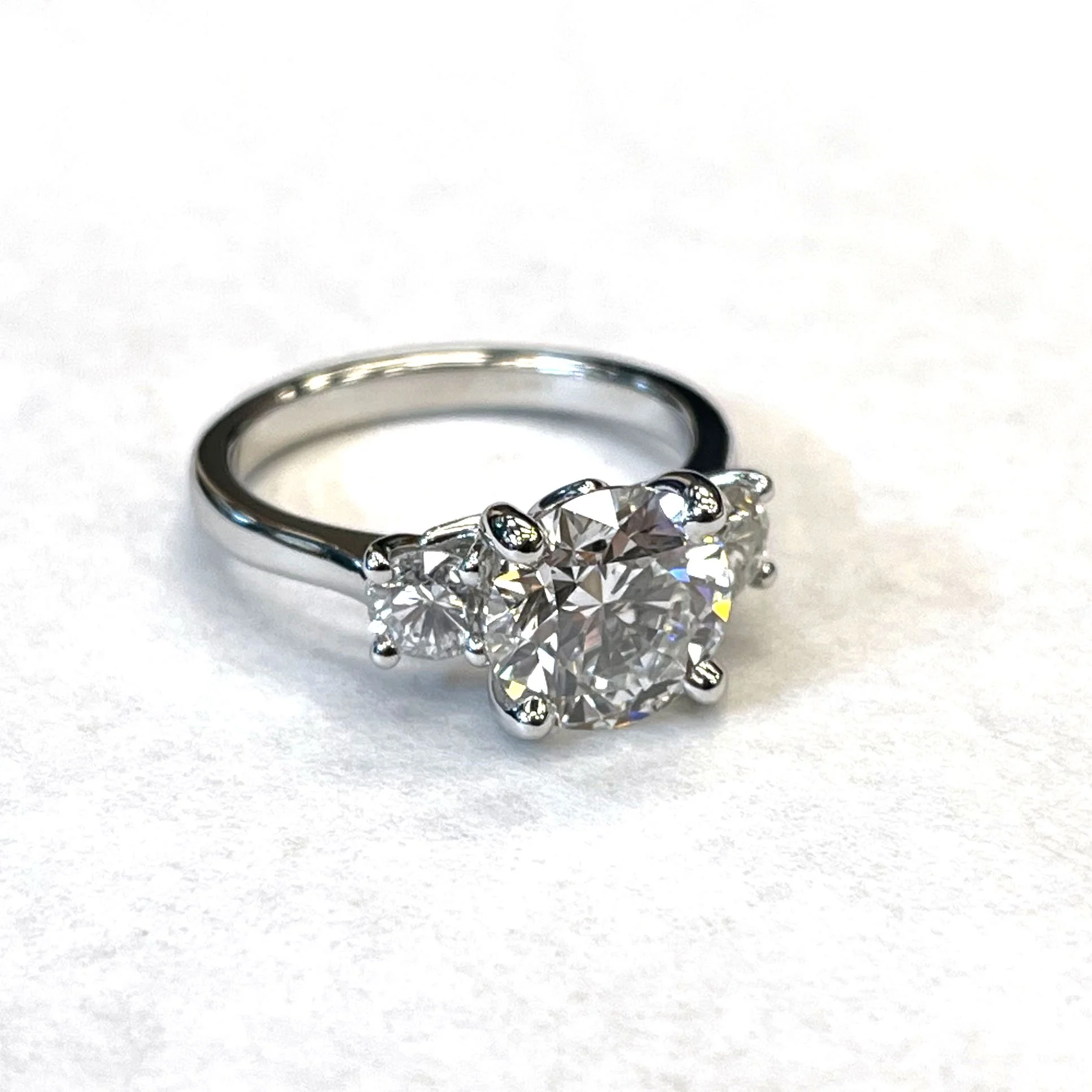 Silver ring with a large heart-shaped diamond in the center and two smaller round diamonds on each side, set against a white background.