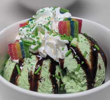 A bowl of mint ice cream topped with whipped cream, rainbow gummy candies, chocolate syrup, and green sprinkles.