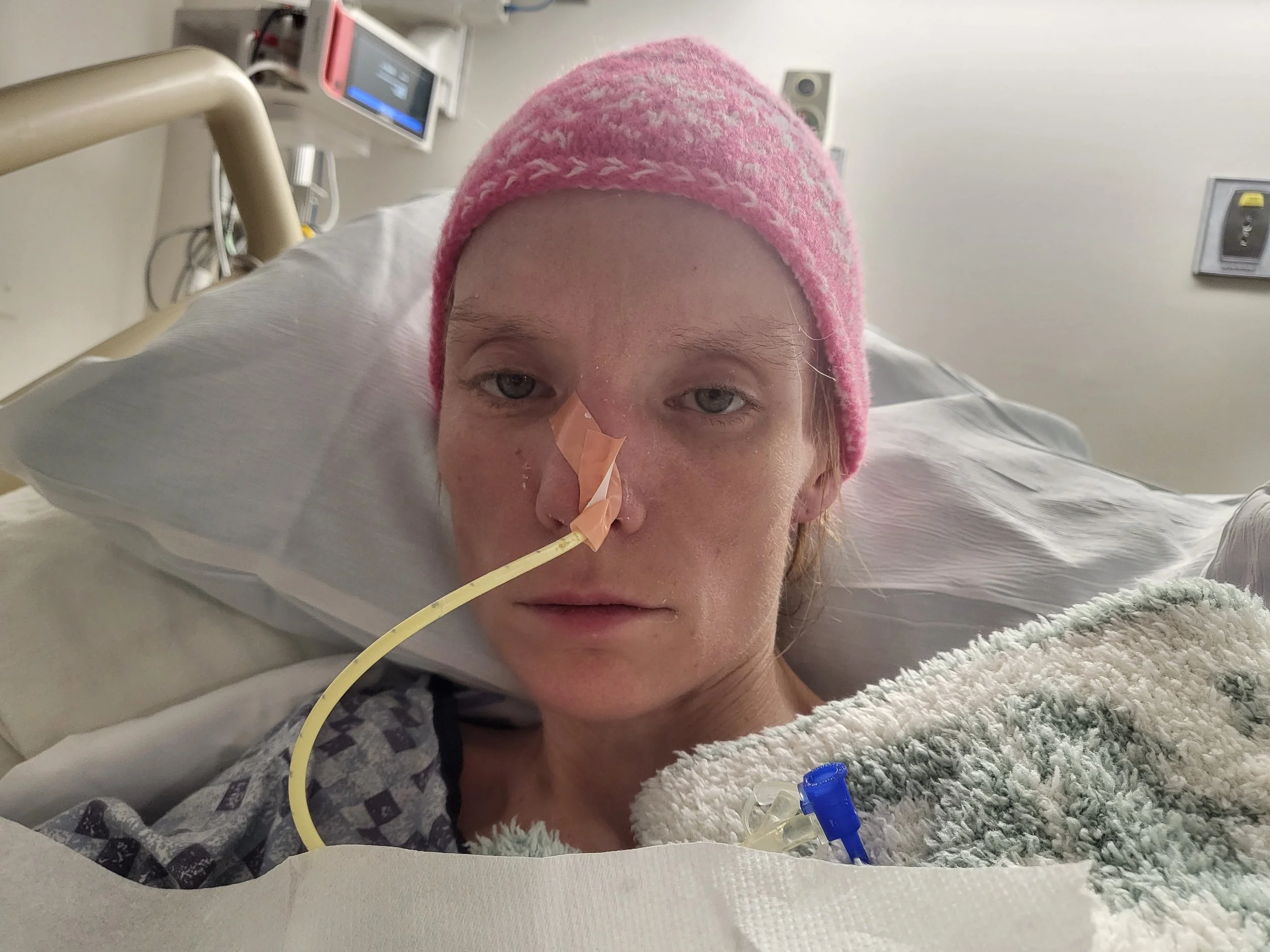 A woman in a hospital bed with a pink knit cap, lying upright with a nasal tube and medical equipment in the background.