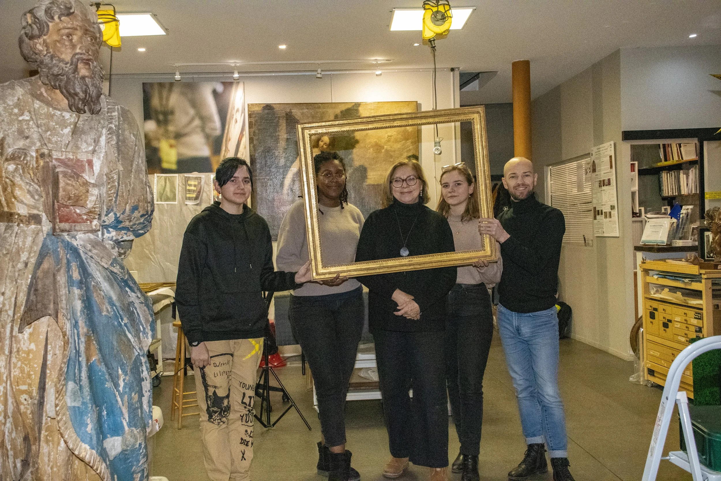 Groupe de cinq personnes posant à l'intérieur d'une galerie ou d'un musée, tenant un cadre doré vide, avec une statue en bois à gauche et des œuvres d'art en arrière-plan.