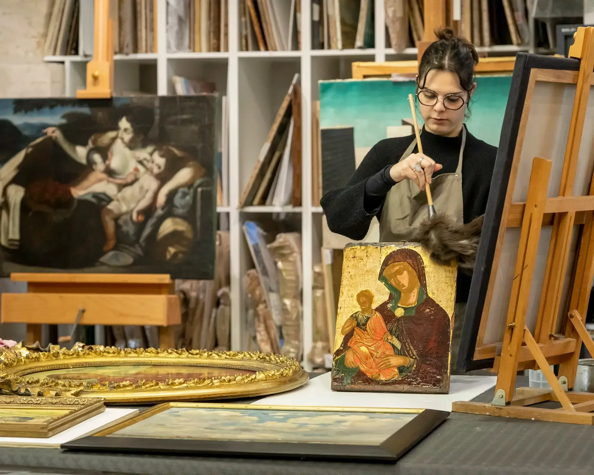 Une femme en train de restaurer une peinture religieuse dans un atelier d'art, entourée de diverses œuvres d'art et d'outils de restauration.