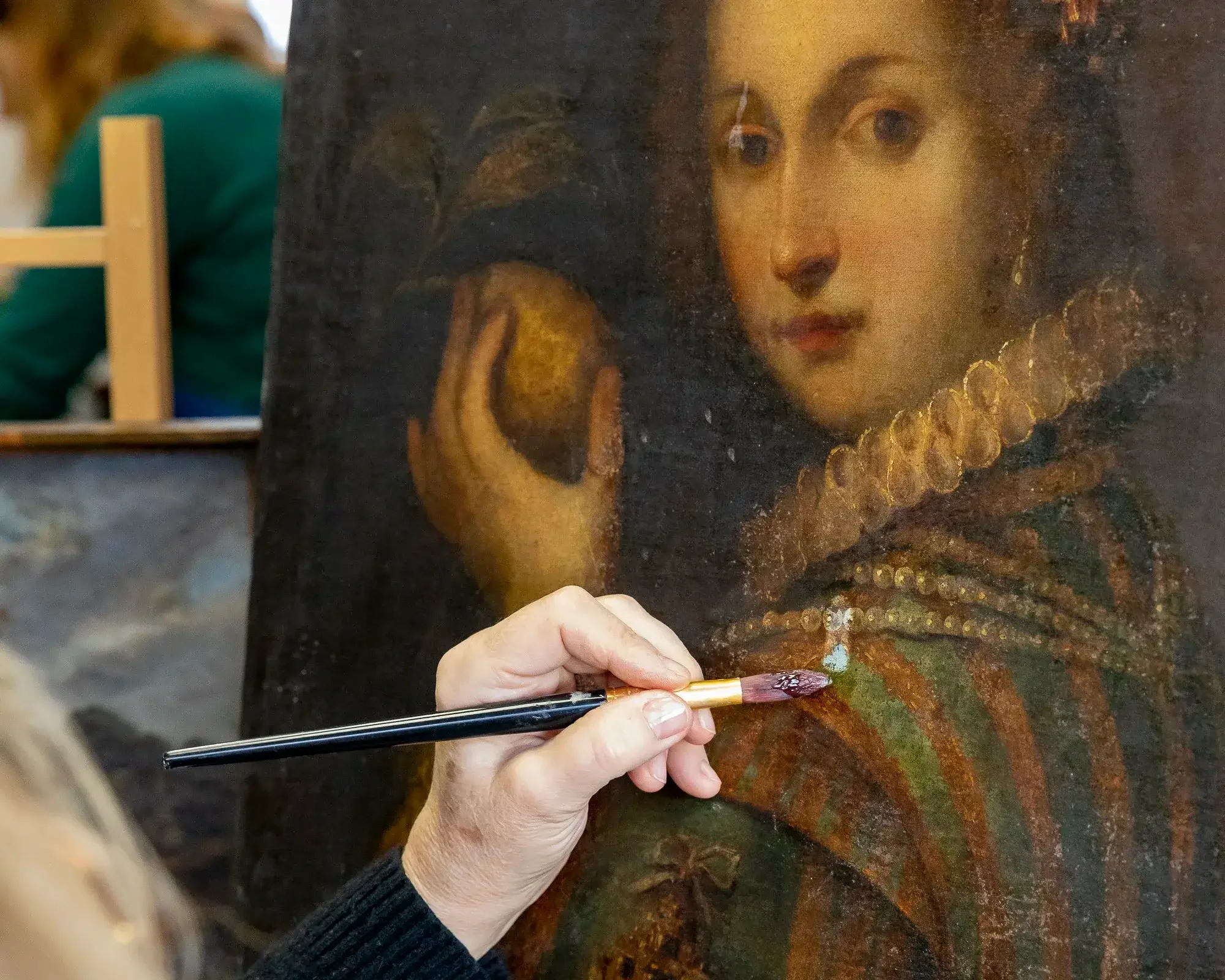 Un restaurateur en train de restaurer une peinture ancienne représentant une femme avec un collier et une robe richement décorée, en utilisant un pinceau fin.