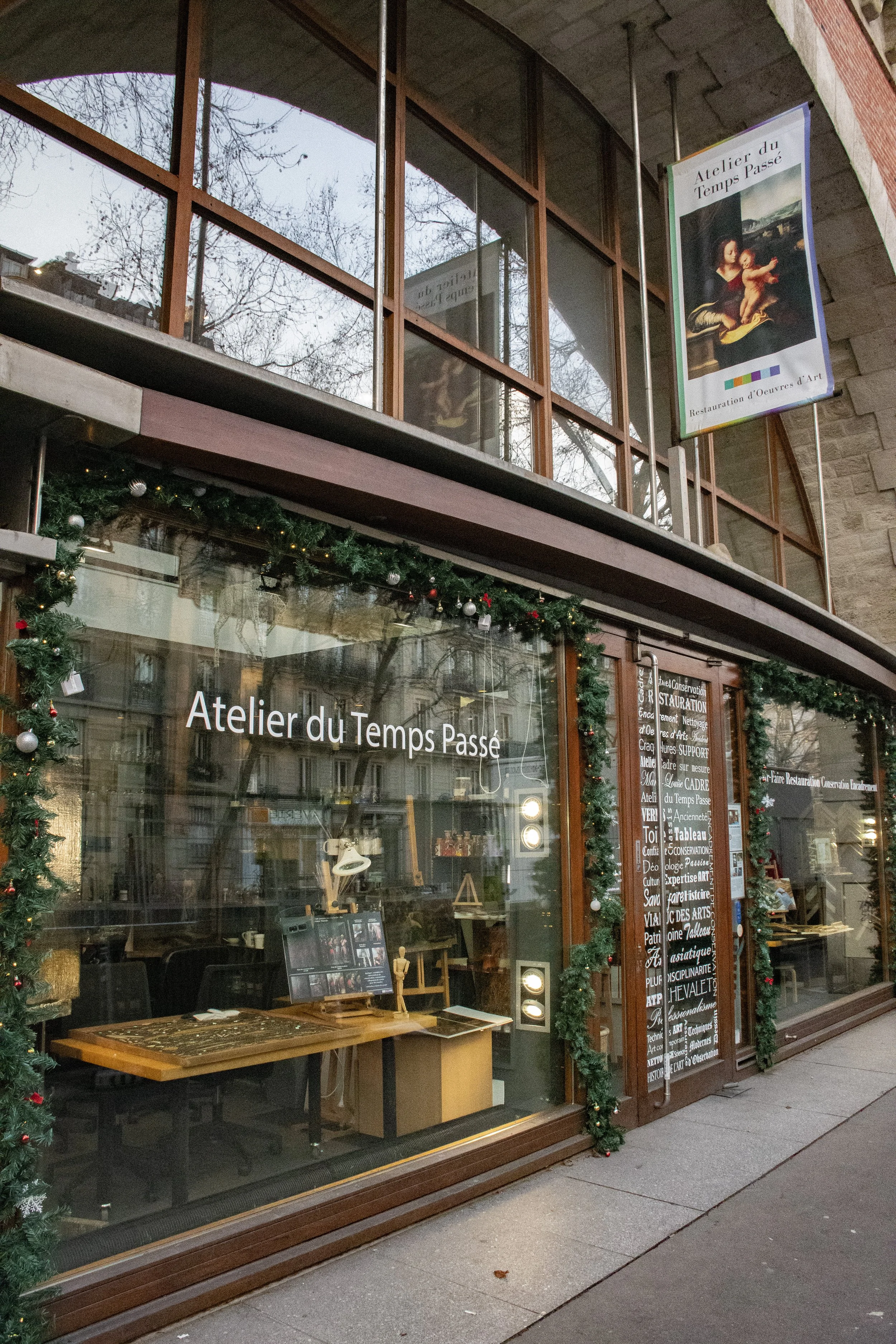 Façade d'un atelier artistique nommé "Atelier du Temps Passé" décorée avec des guirlandes de Noël, vitres avec affiches et objets d'art à l'intérieur.