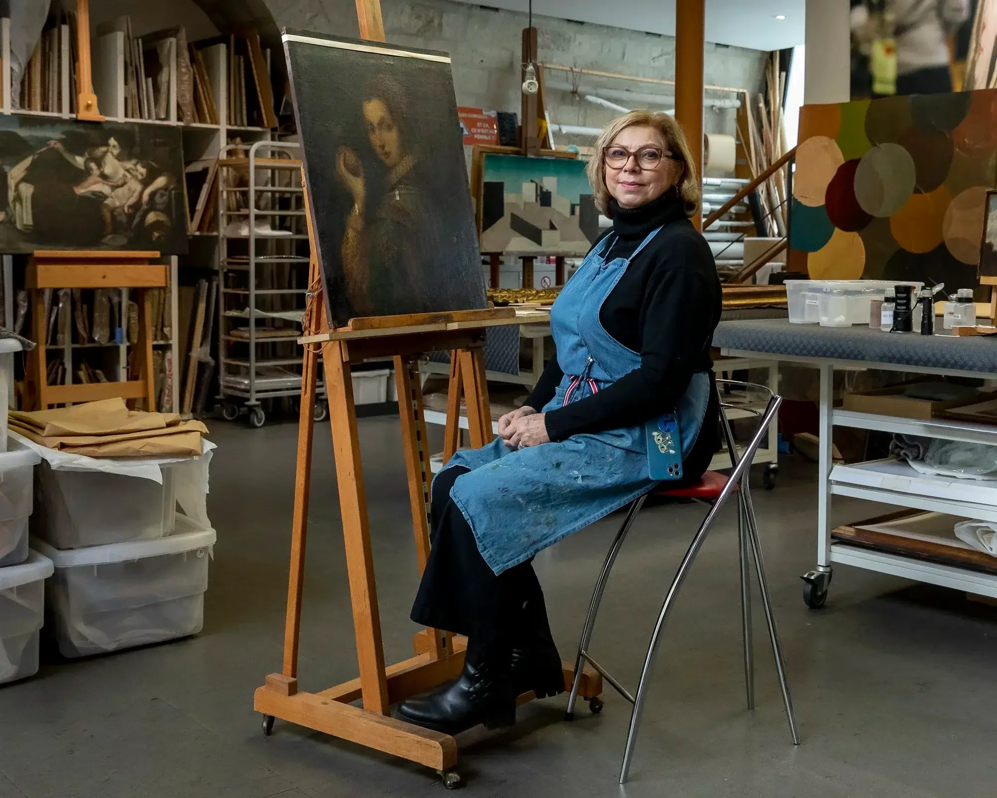Une femme assise dans un atelier d'art, portant un tablier bleu, devant une toile. L'atelier est rempli de tableaux et de matériaux artistiques.