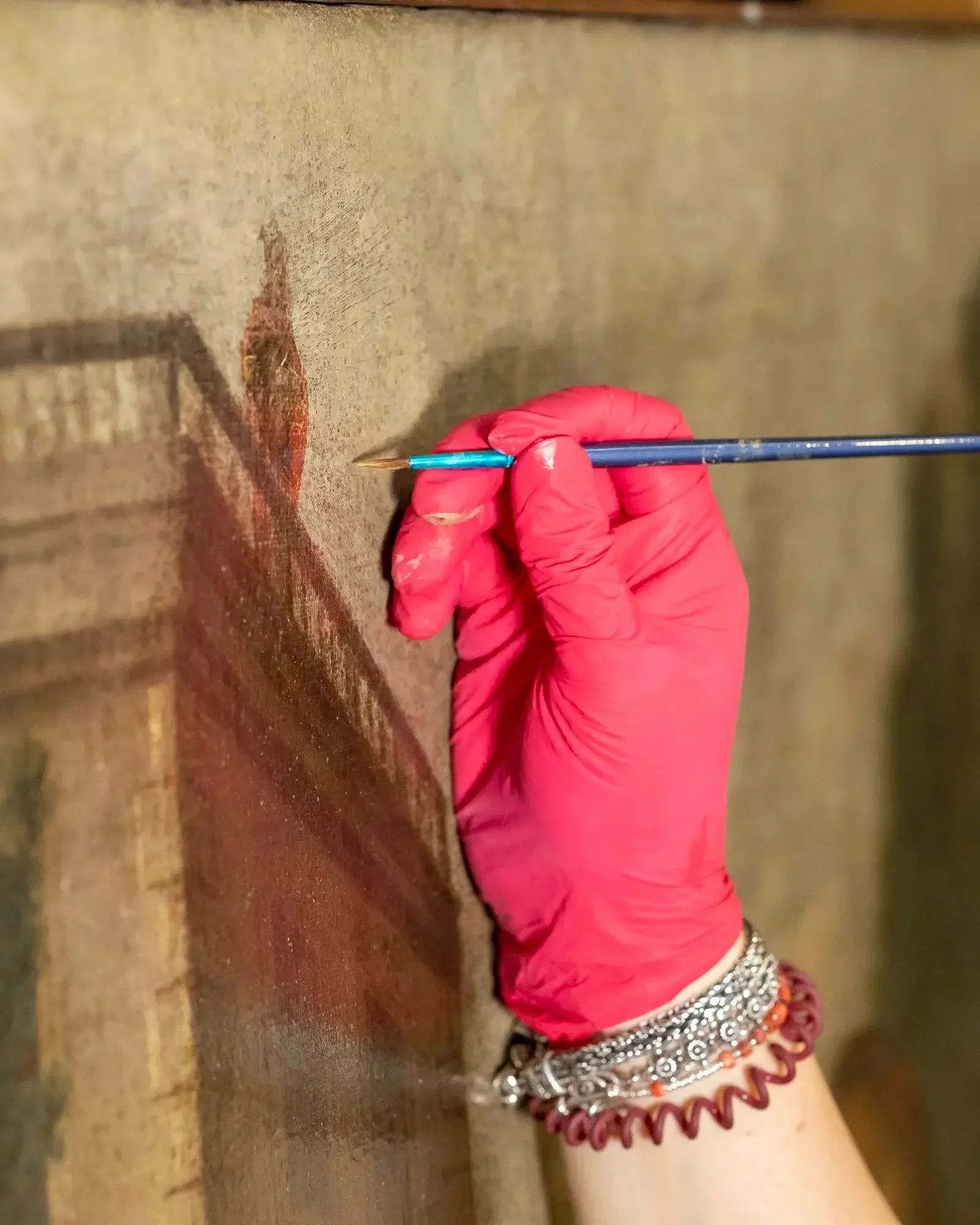 Une main portant un gant rose tient un pinceau fin et travaille sur une toile ou une surface avec une teinte rouge ou marron.