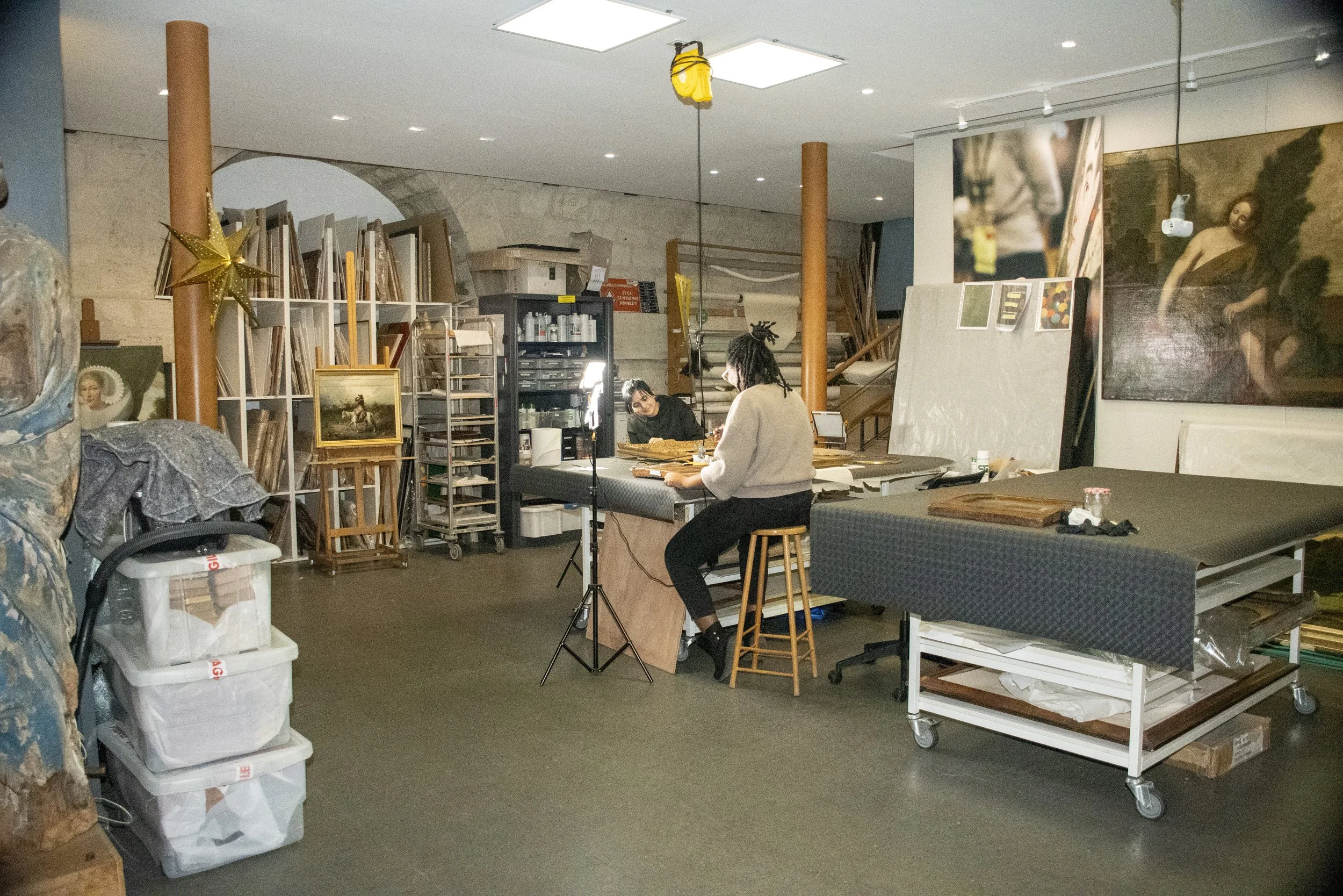 Deux personnes travaillent dans un atelier d'art, entourées de toiles, de cadres, et de matériaux divers avec une lumière dirigée sur leur espace de travail.