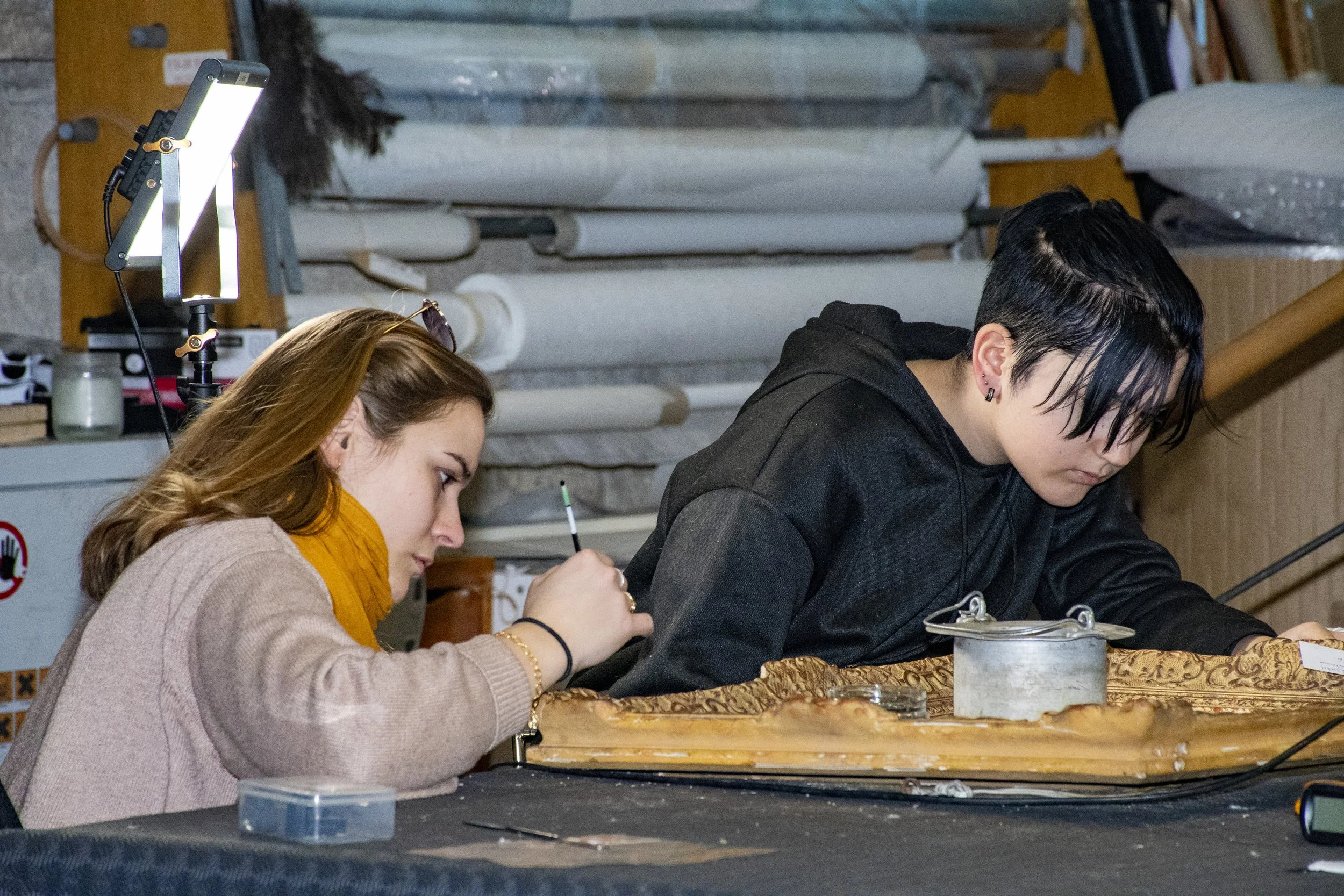 Deux jeunes femmes travaillent sur un projet artistique ou de restauration, l'une avec un pinceau et l'autre concentrée sur un objet ancien sur une table.