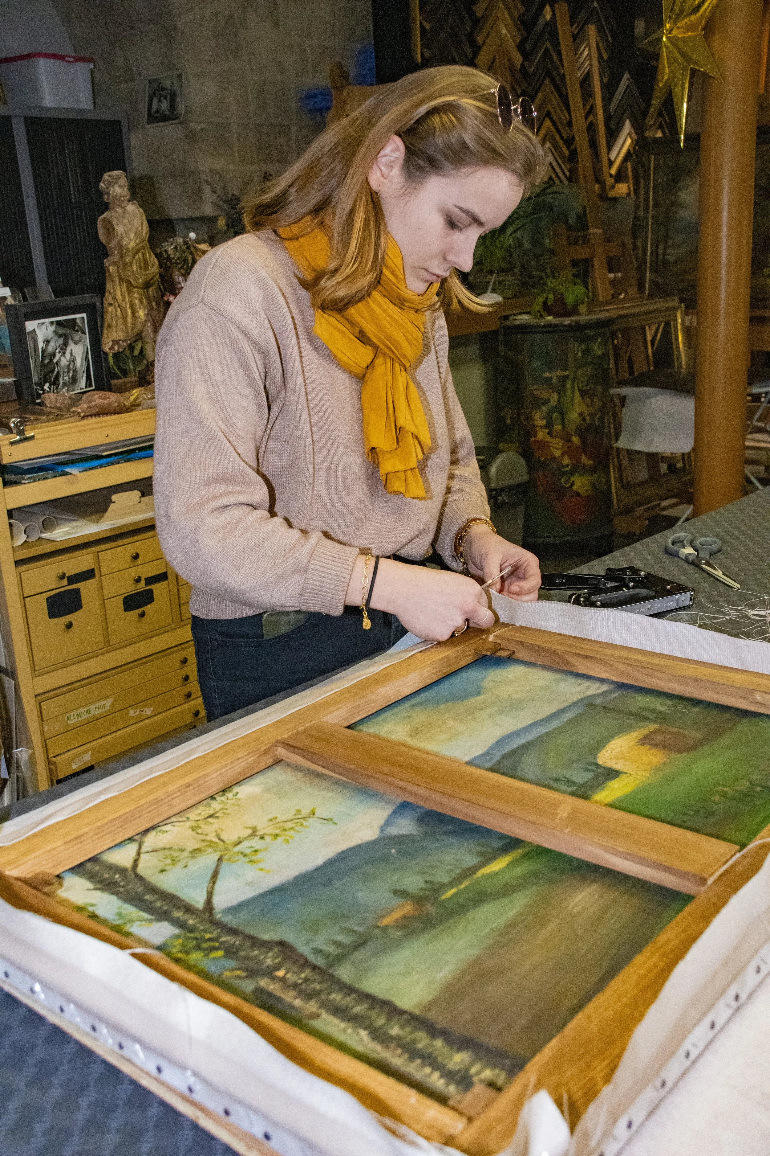 Femme en train d'encadrer une peinture à l'huile représentant un paysage de campagne avec des montagnes, un arbre, un champ et un ciel bleu, dans un atelier d'art.
