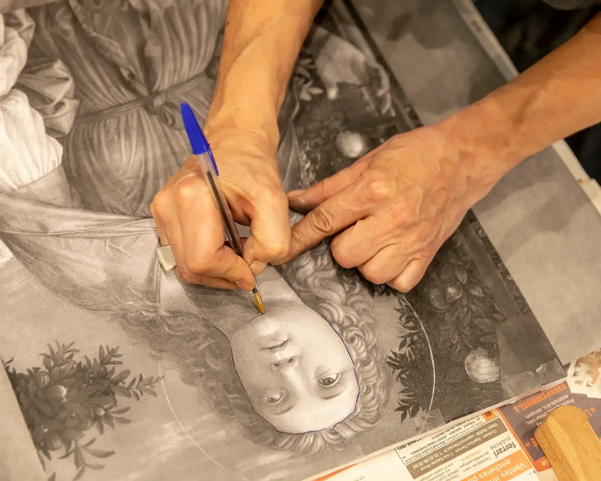 Une personne utilise un stylo pour dessiner ou graver un portrait en noir et blanc d'une femme avec des cheveux bouclés, entourée de plantes et de fruits, sur une surface en papier ou en toile.