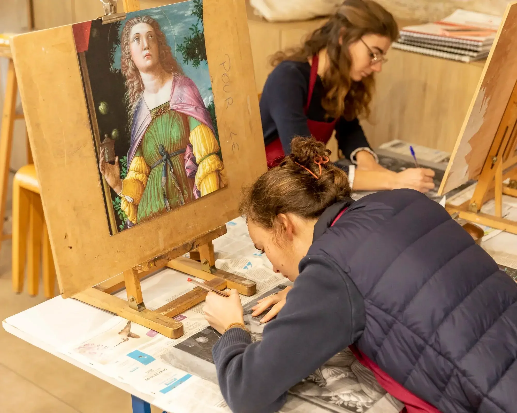 Deux jeunes femmes restaurent une peinture ancienne représentant une femme avec des vêtements colorés, dans un atelier.