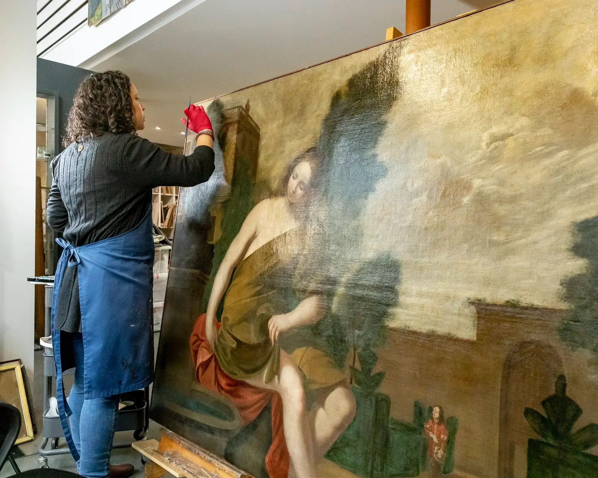 Une femme restauratrice en train de réparer un tableau ancien représentant une femme assise, dans un atelier d'art.