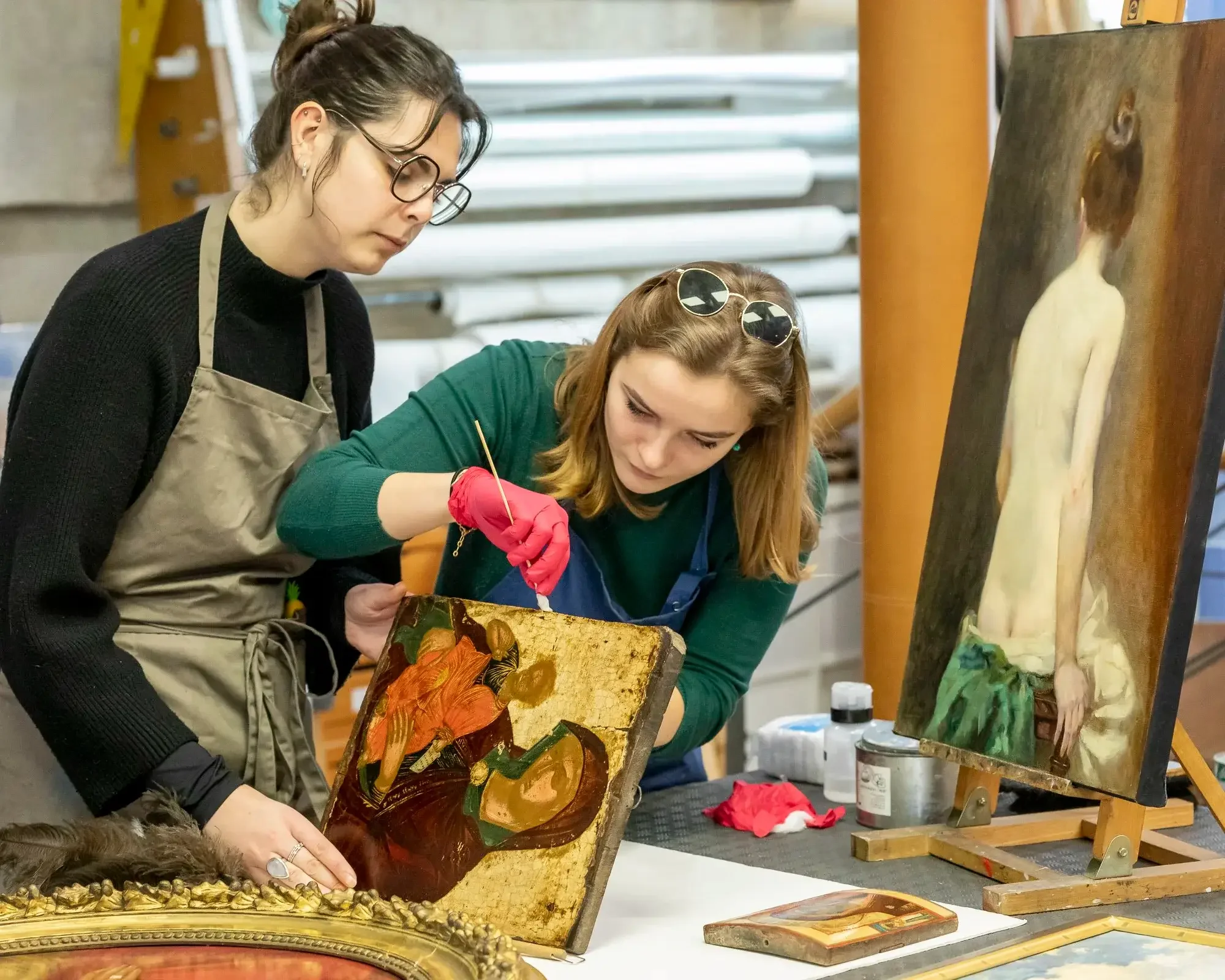Deux femmes restaurent et nettoient des icônes ou des peintures d'art religieux dans un atelier. L'une d'elles tient une pince et l'autre une pièce de sécurité ou un outil, sous l'œil attentif d'une première femme.