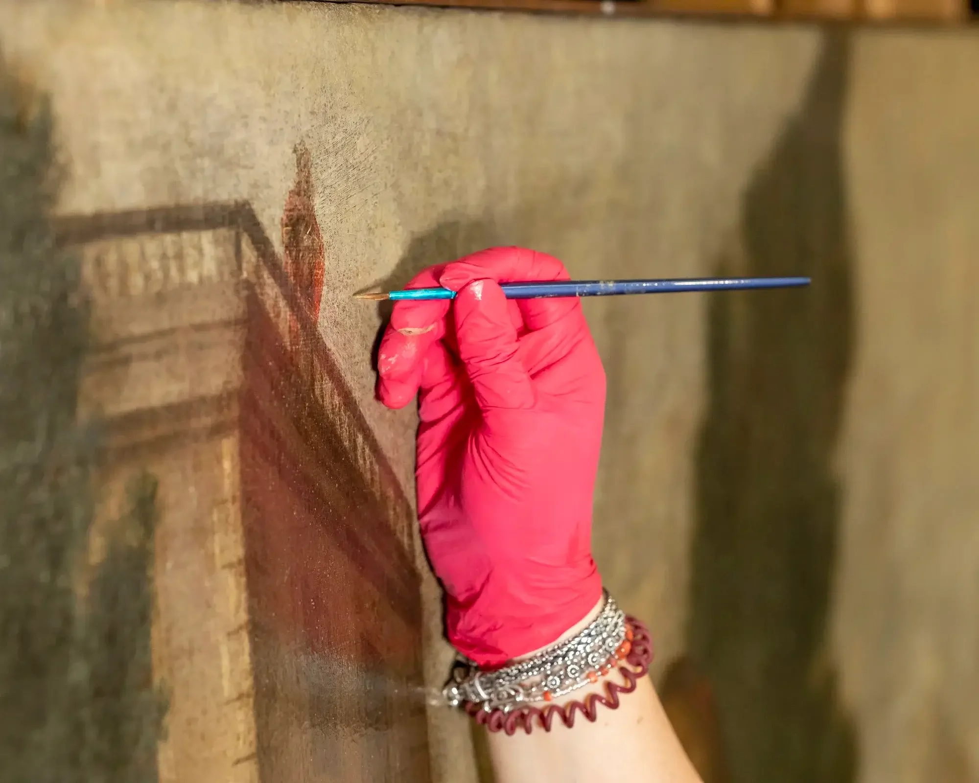Main tenant un pinceau fin pour restaurer une peinture ancienne. La main porte un gant rose et porte plusieurs bracelets en acier et en caoutchouc.