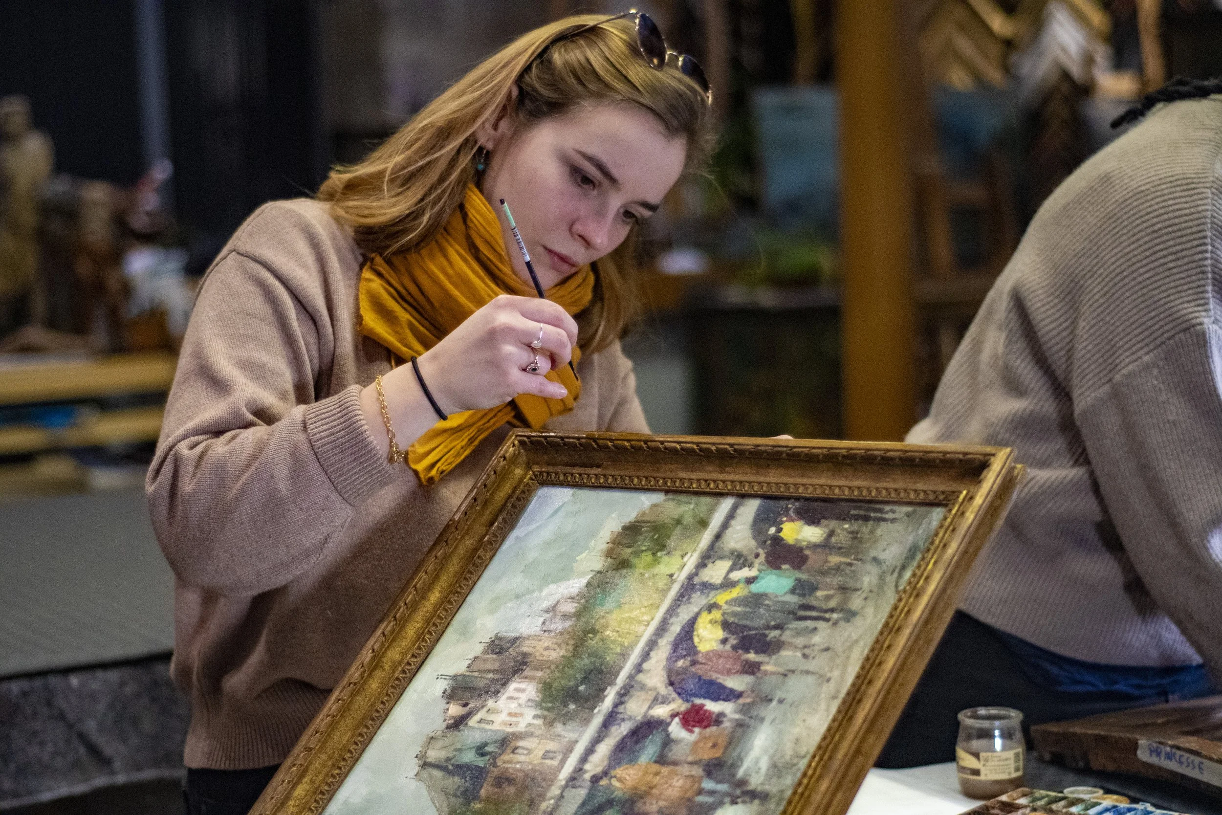 Une jeune femme avec une écharpe orange et des lunettes de soleil sur la tête, en train de peindre ou restaurer un tableau dans un atelier.