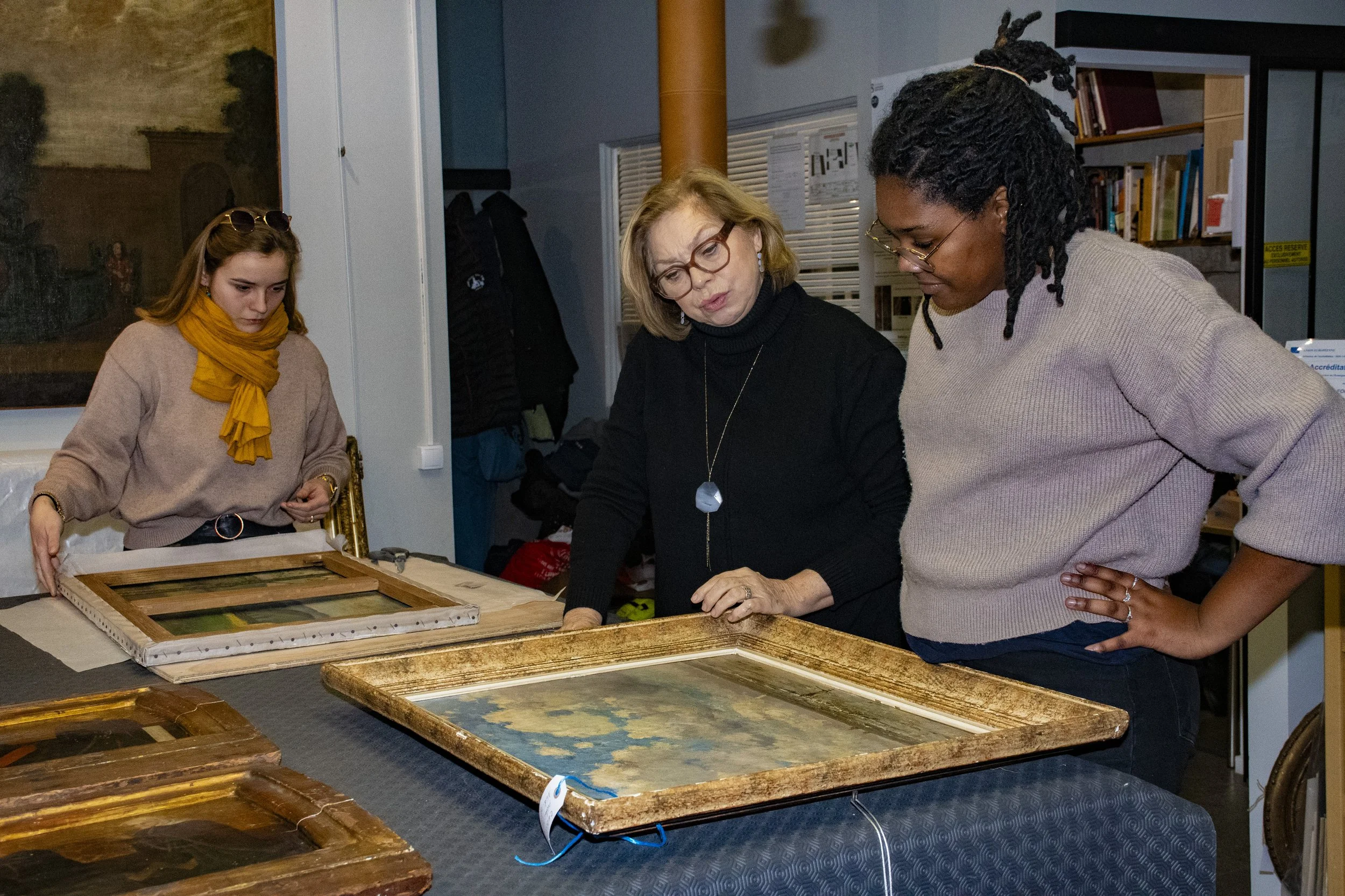 Trois femmes regardent et discutent autour de cadres de peinture ou de dessin dans une salle, avec des livres et des objets en arrière-plan.