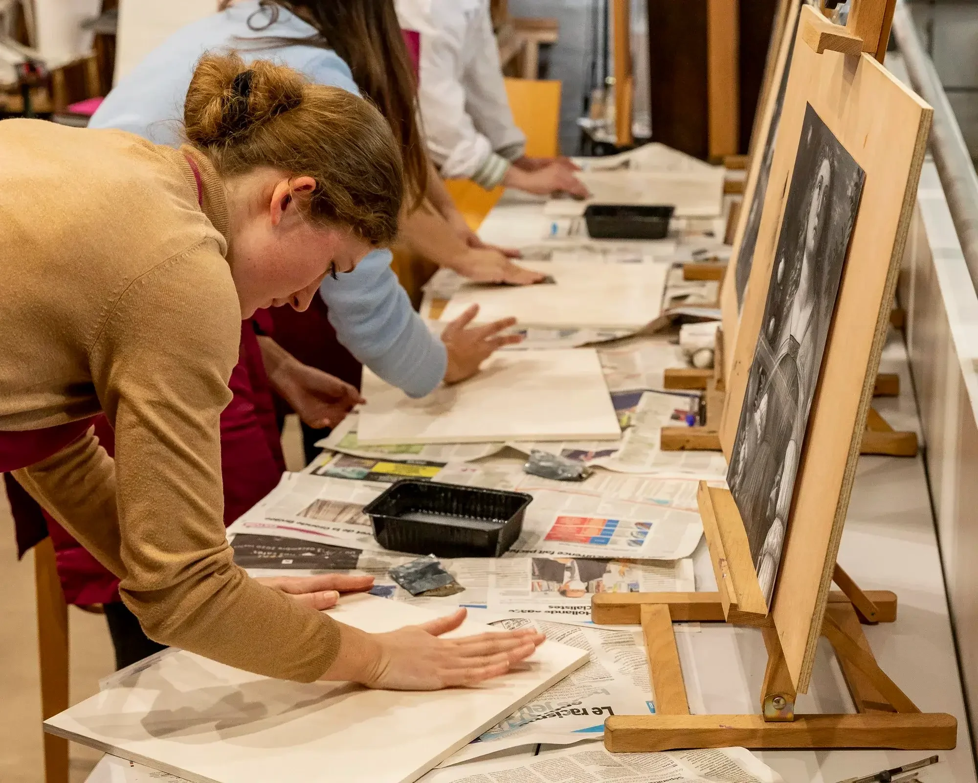 Groupe de personnes participant à une activité artistique ou artisanale, probablement une peinture ou une sculpture, dans un atelier, en train de travailler sur leurs œuvres avec des pinceaux et des matériaux divers, avec des toiles ou des supports e