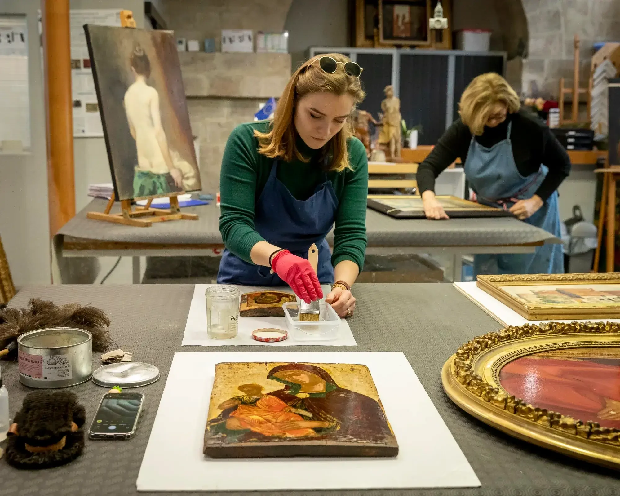 Deux femmes restaurent ou restaurent des œuvres d'art dans un atelier, avec des peintures et des cadres anciens sur la table.