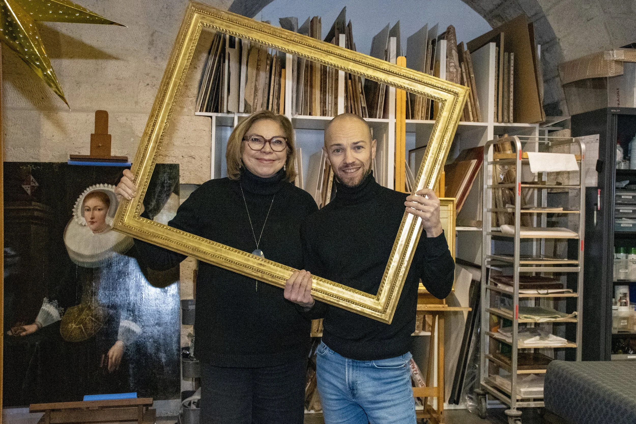 Deux personnes tenant un cadre doré dans un atelier avec des étagères remplies de livres et œuvres d'art