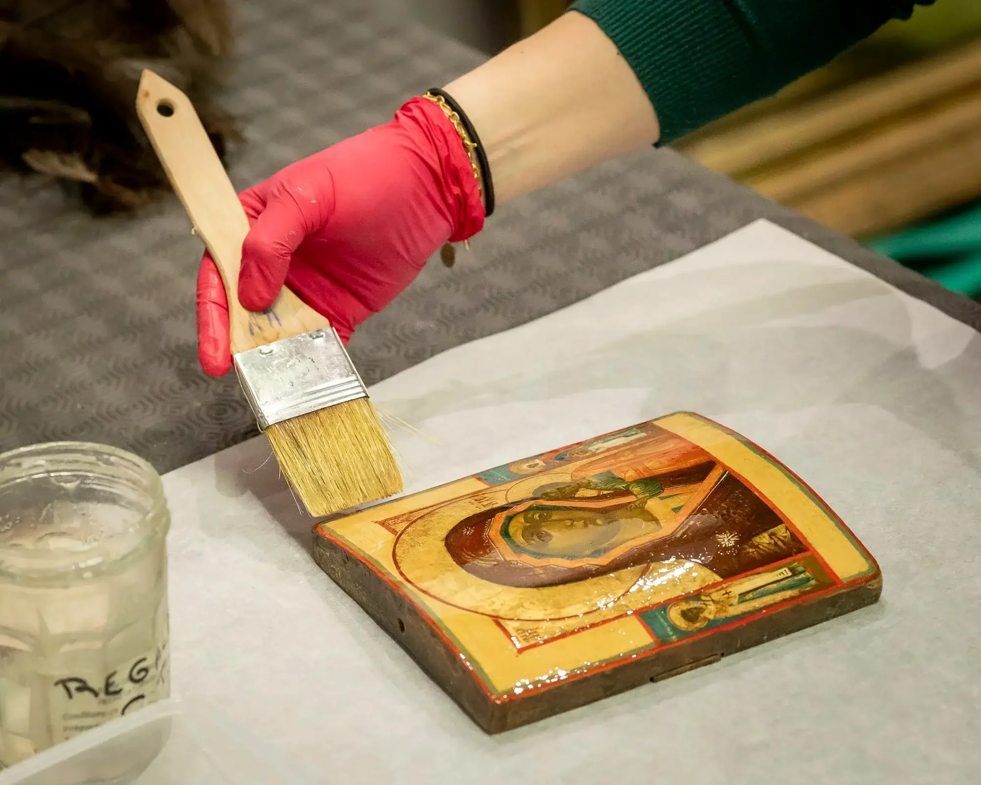 Une personne portant un gant rouge nettoie une icône religieuse avec un pinceau large. L'icône représente probablement une figure religieuse et est posée sur une surface. Un pot de verre est visible à côté.