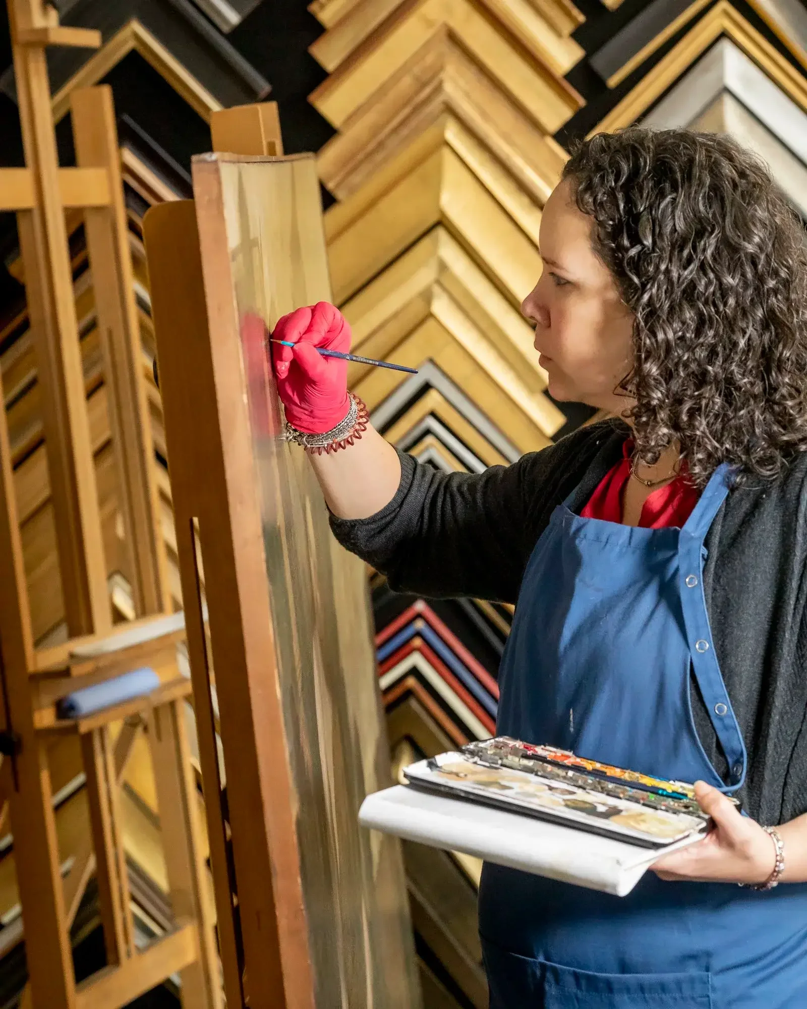 Une femme avec des cheveux bouclés, portant un tablier bleu, peint une toile avec un pinceau dans un magasin de cadres. Elle tient une palette de peintures en accompagnement.