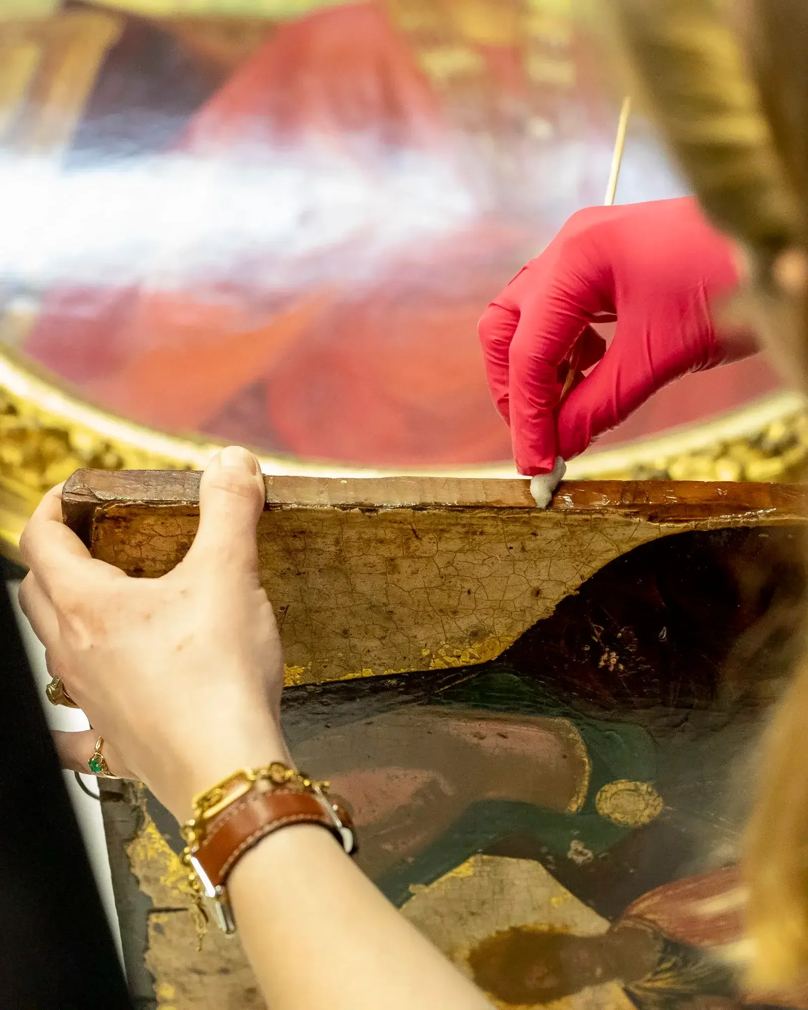 Une personne portant des gants rouges nettoie une peinture ancienne avec un outil pointu.