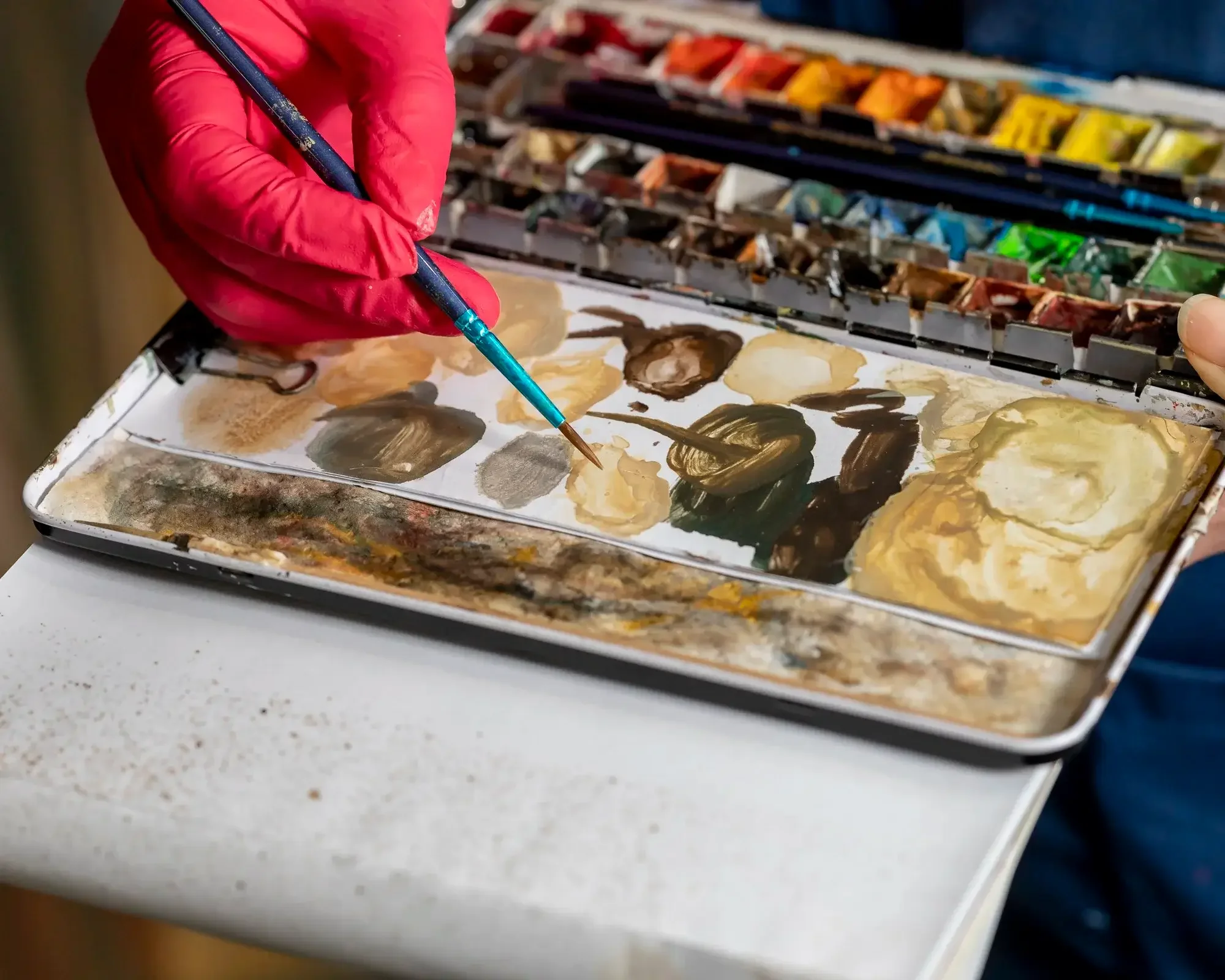 Une personne portant un gant rouge utilise un pinceau pour mélanger des couleurs dans une palette d'aquarelle, avec des tubes de peinture colorée visibles en haut.