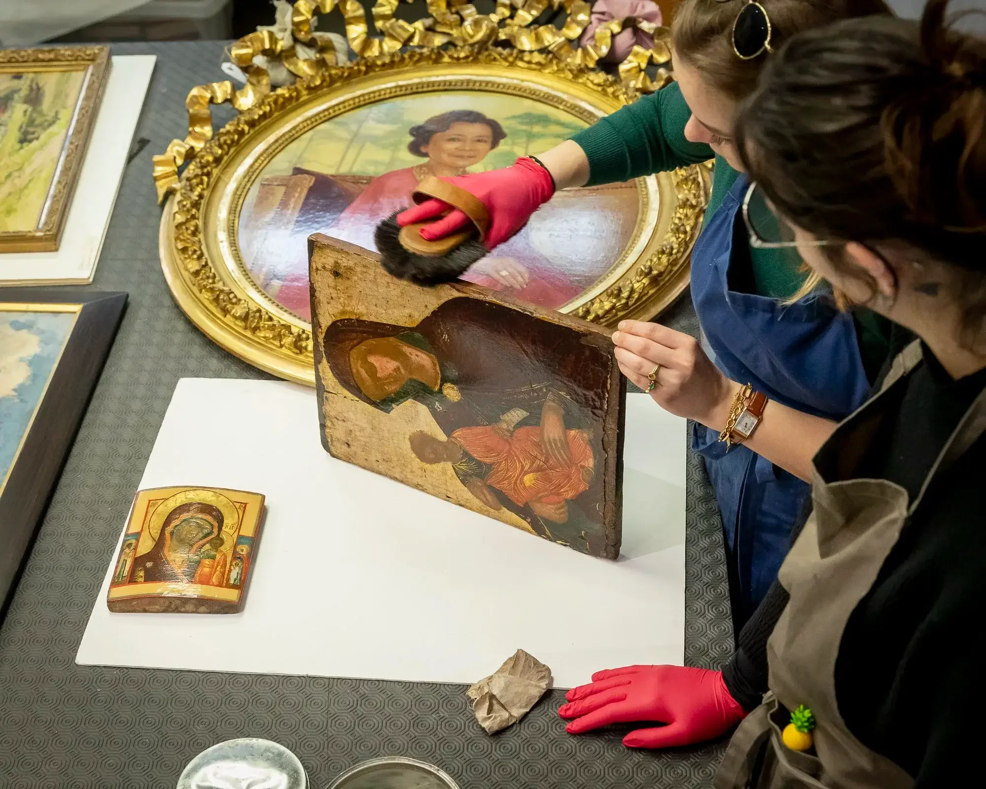 Deux personnes nettoient une ancienne peinture religieuse derrière un cadre doré. Il y a d'autres œuvres d'art visibles sur la table.