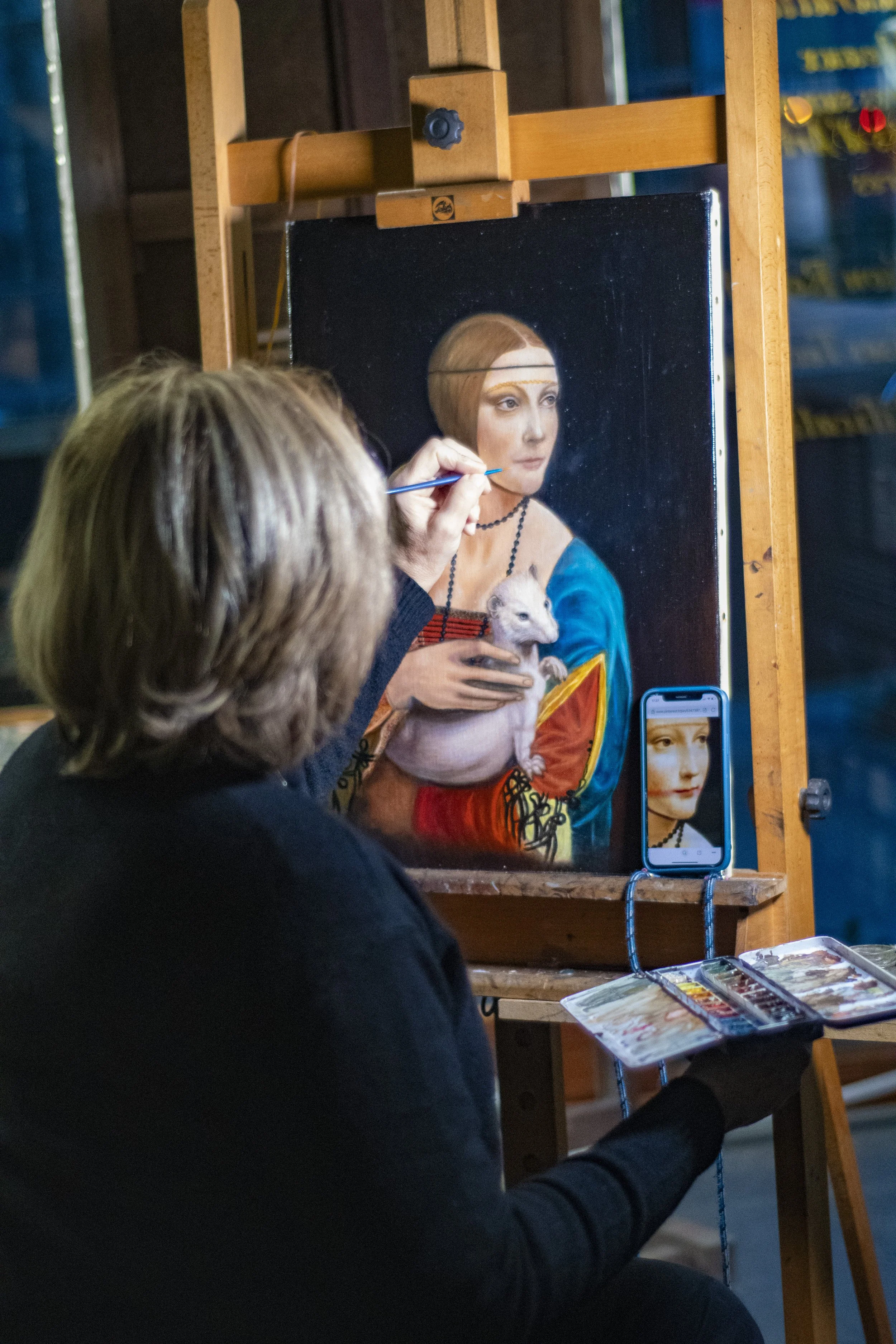 Un artiste peint un tableau représentant une femme avec une tête rousse tenant un rat blanc, tout en utilisant un téléphone pour prendre une photo de son œuvre.