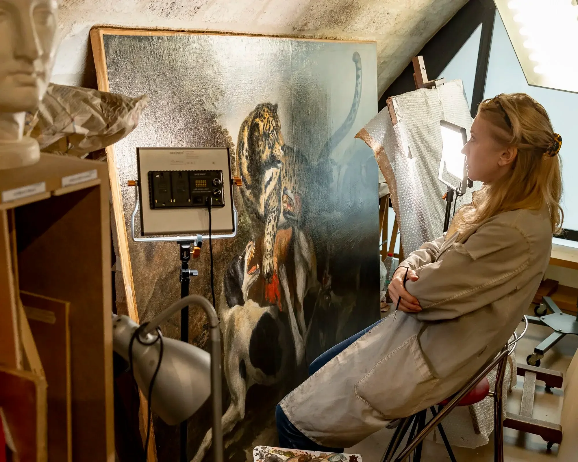 Une femme est assise à côté d'une grande peinture de tigre. Elle regarde le tableau, avec des lumières d'éclairage autour, dans un atelier d'art.