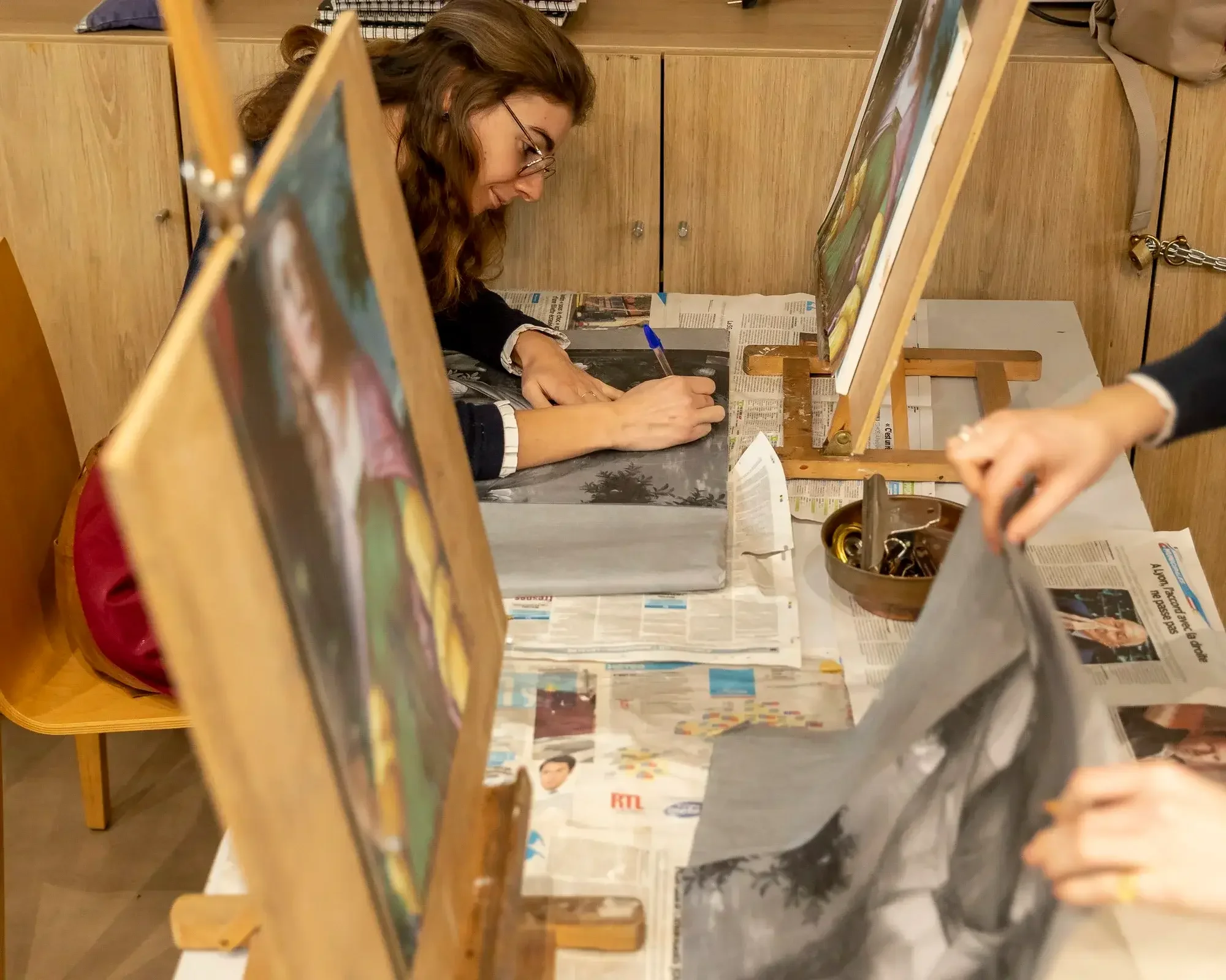 Une femme peint sur une toile, sous un étroit cadre en bois, dans un espace de création artistique. Elle est concentrée et utilise un pinceau, avec d'autres personnes qui participent également à la peinture autour d'elle.